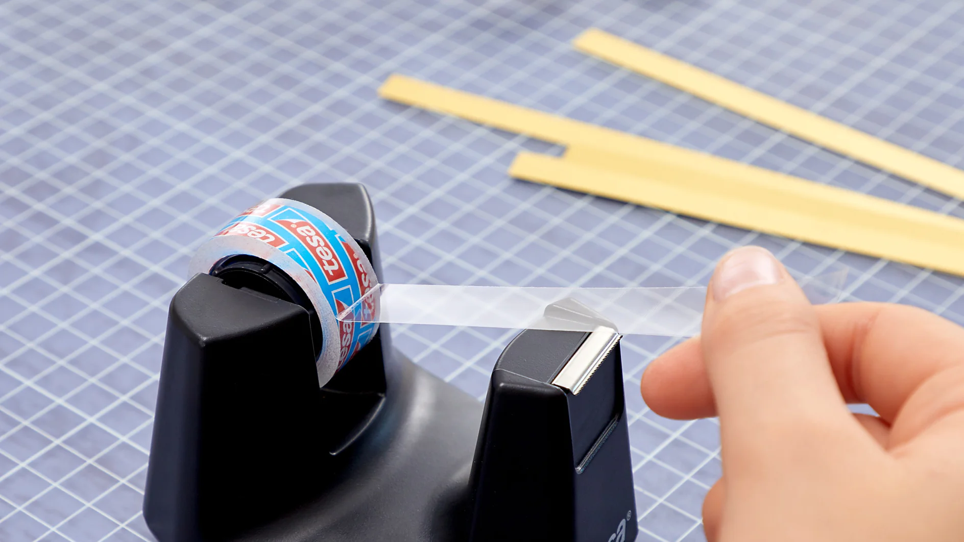 A hand pulls clear tesa tape from a black tape dispenser marked tesa on a chequered cutting mat. Yellow strips and a decorative paper design are visible in the background. (This text has been generated by AI)