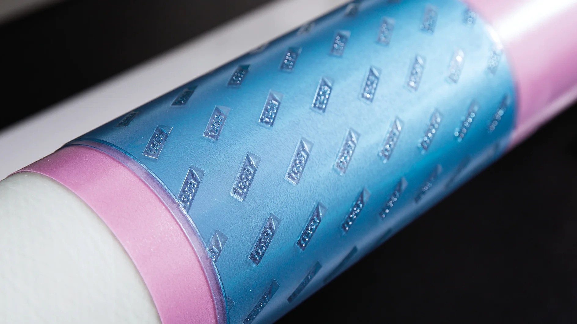 Close-up of a cylindrical object wrapped in light blue and pink material featuring a textured diamond pattern. The surface has a glossy finish, and the background is out of focus, displaying contrasting light and dark areas. (This text has been generated by AI)
