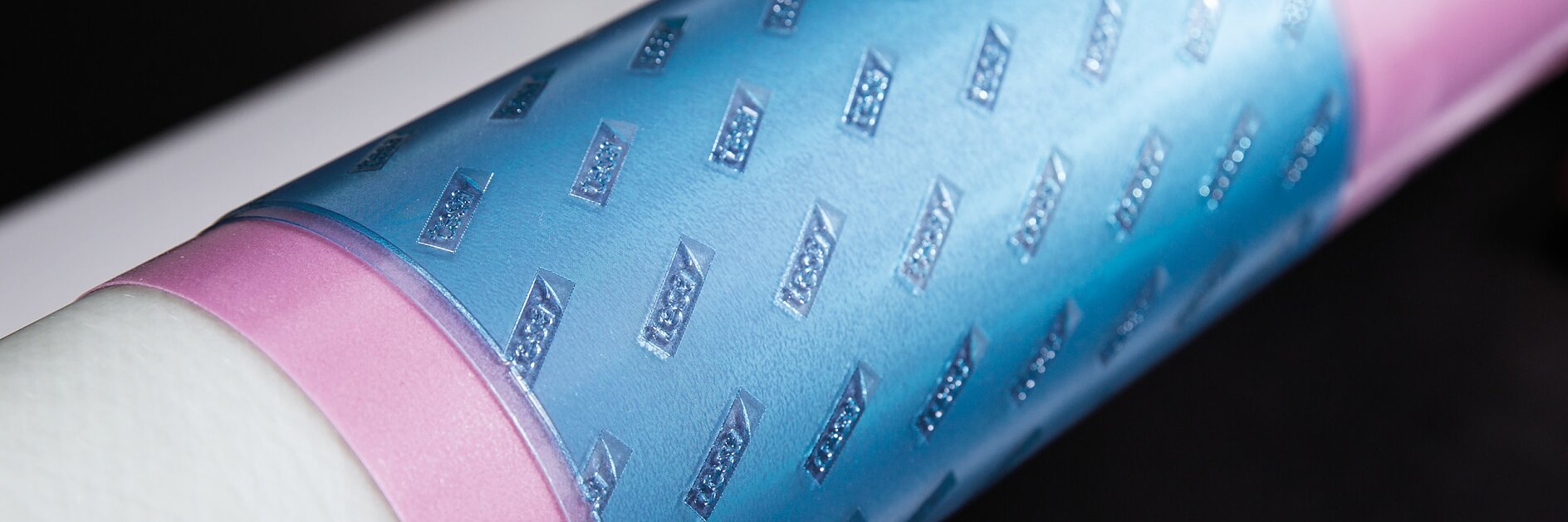 Close-up of a cylindrical object wrapped in light blue and pink material featuring a textured diamond pattern. The surface has a glossy finish, and the background is out of focus, displaying contrasting light and dark areas. (This text has been generated by AI)