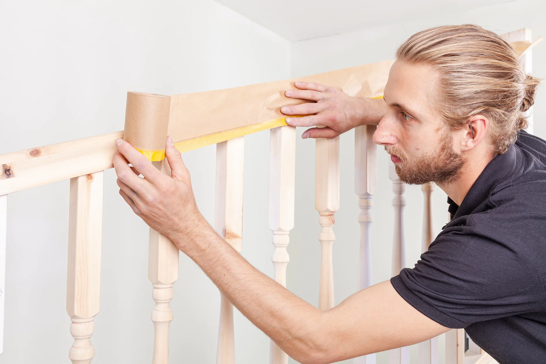 A man with a beard and tied-back hair applies tesa tape to a wooden staircase railing. He is focused on securing the tesa tape along the top edge of the balusters, preparing the railing for painting or staining. (This text has been generated by AI)