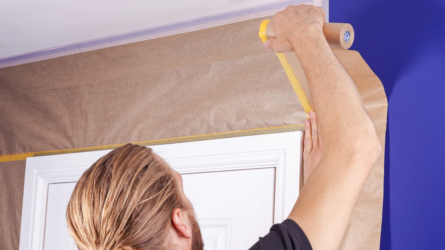 A person with long hair in a bun is applying tesa tape to a brown paper covering around a door frame. The wall is painted blue. They are wearing a black shirt and holding the tesa tape in their right hand. (This text has been generated by AI)