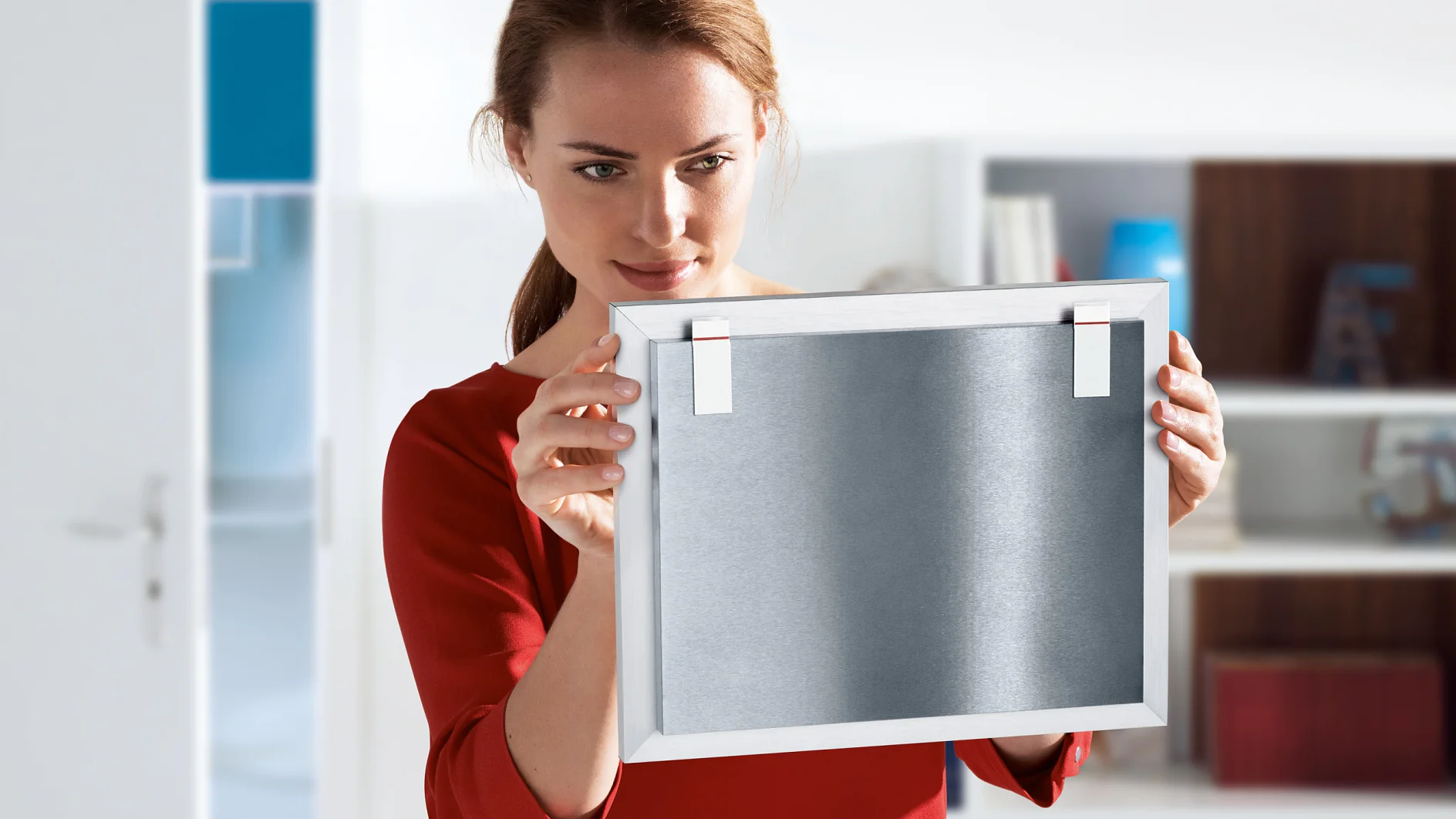 A woman in a red shirt examines a framed metal object, holding it in both hands. She stands in a brightly lit room with a bookshelf in the background and an open door to the left. The frame is secured with tesa tape, offering reliable and sturdy support for her display. (This text has been generated by AI)