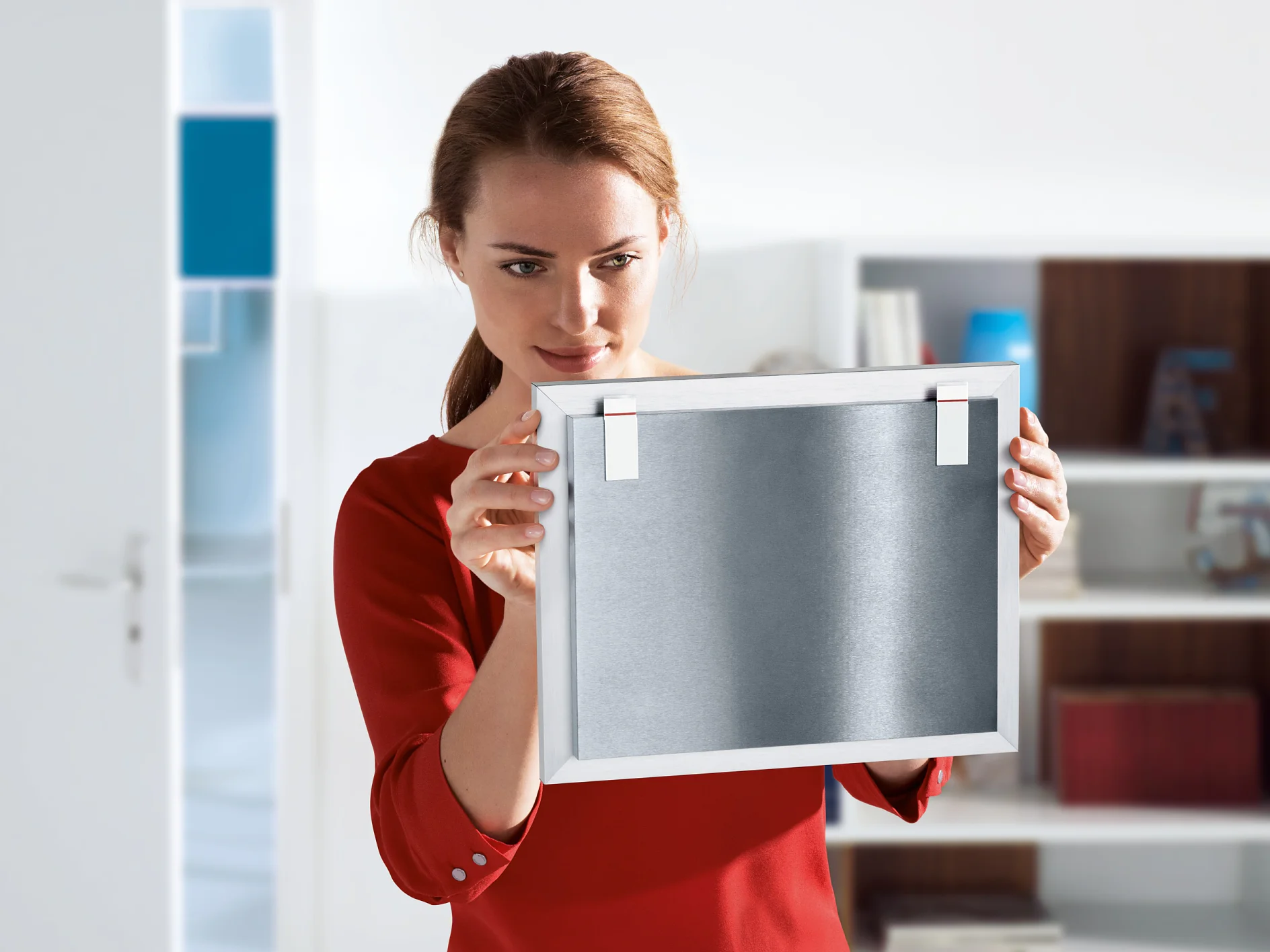 A woman in a red shirt examines a framed metal object, holding it in both hands. She stands in a brightly lit room with a bookshelf in the background and an open door to the left. The frame is secured with tesa tape, offering reliable and sturdy support for her display. (This text has been generated by AI)