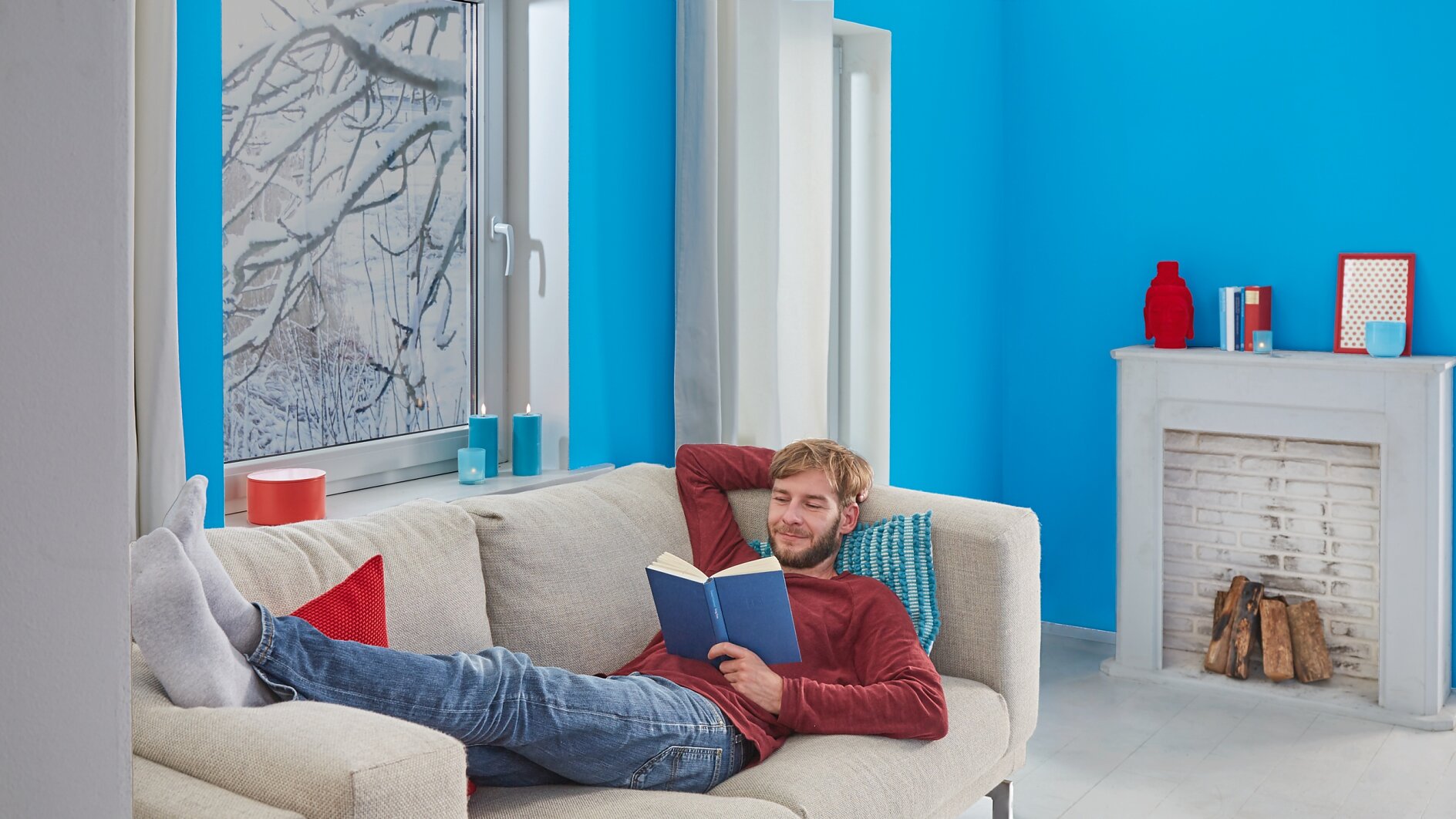 A man is relaxing on a beige couch, reading a book in a room with bright blue walls. He wears a red sweater and jeans, with his legs resting on the couch. In the background, theres a white fireplace and a window showcasing snowy branches. (This text has been generated by AI)