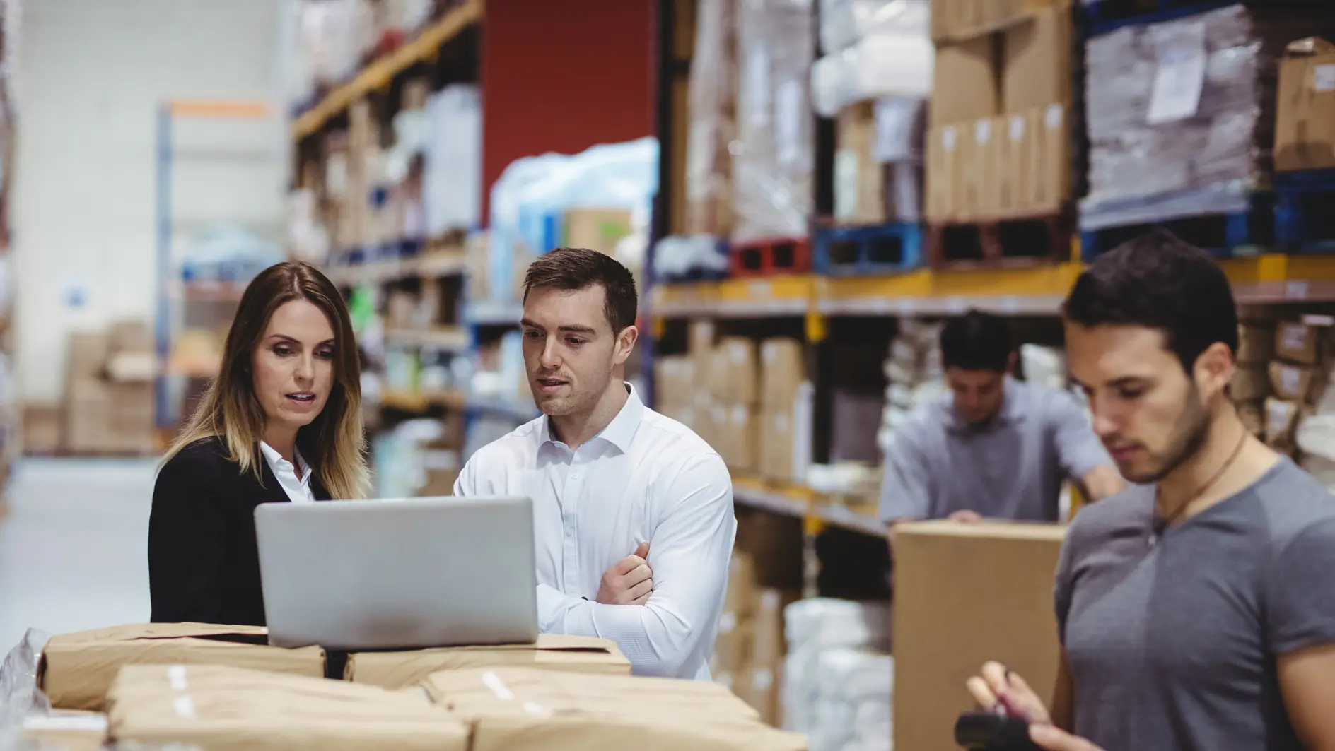 A group of warehouse workers stand by a table filled with packages. Two are engaged with a laptop, while another tallies items using a handheld device. Shelves stocked with boxes and supplies, including tesa tape, are visible in the background. (This text has been generated by AI)