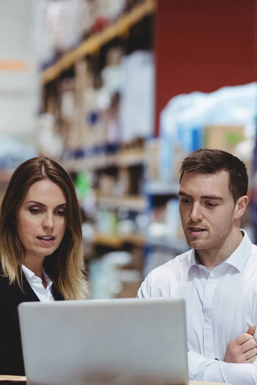 A group of warehouse workers stand by a table filled with packages. Two are engaged with a laptop, while another tallies items using a handheld device. Shelves stocked with boxes and supplies, including tesa tape, are visible in the background. (This text has been generated by AI)