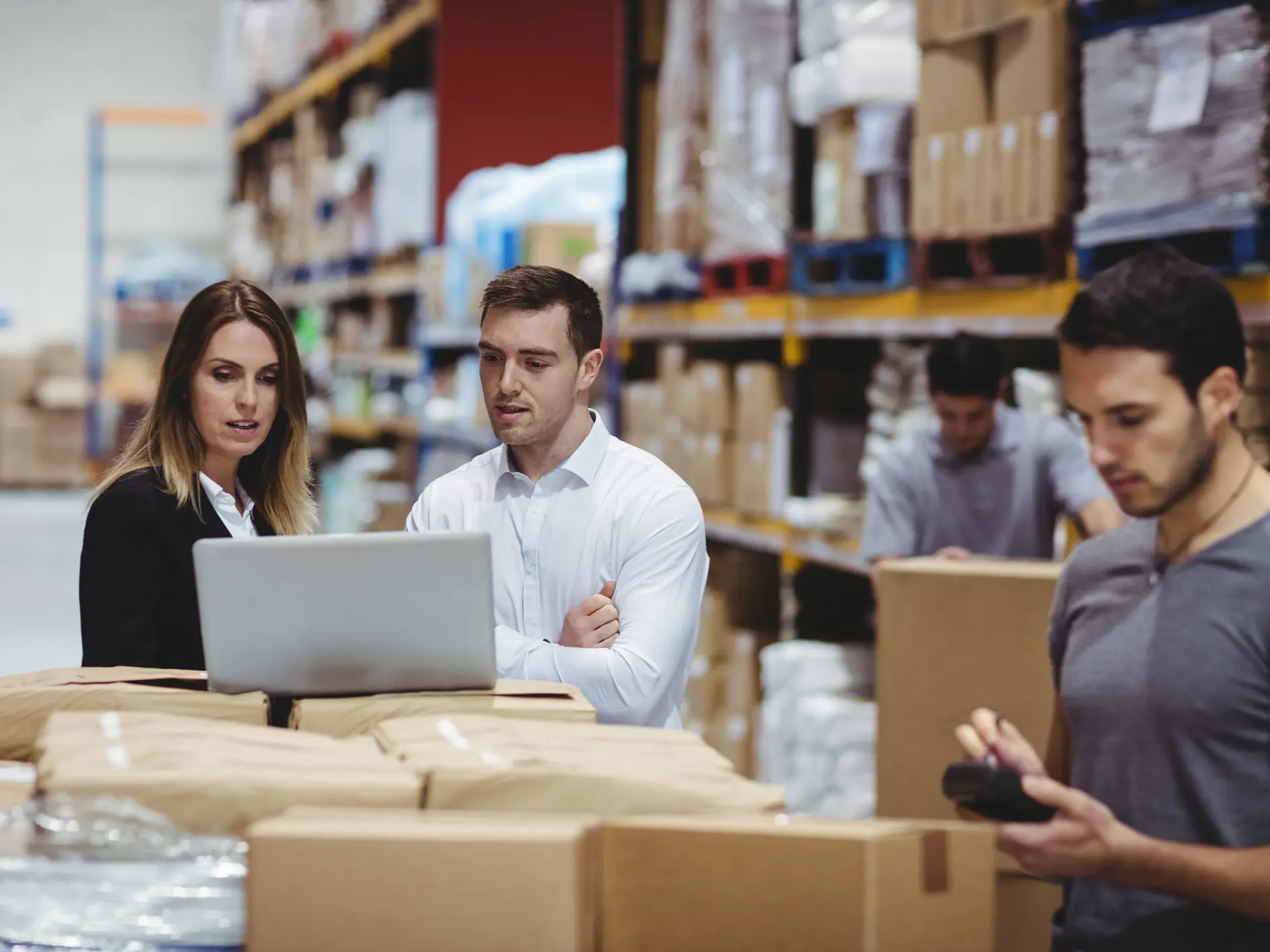A group of warehouse workers stand by a table filled with packages. Two are engaged with a laptop, while another tallies items using a handheld device. Shelves stocked with boxes and supplies, including tesa tape, are visible in the background.