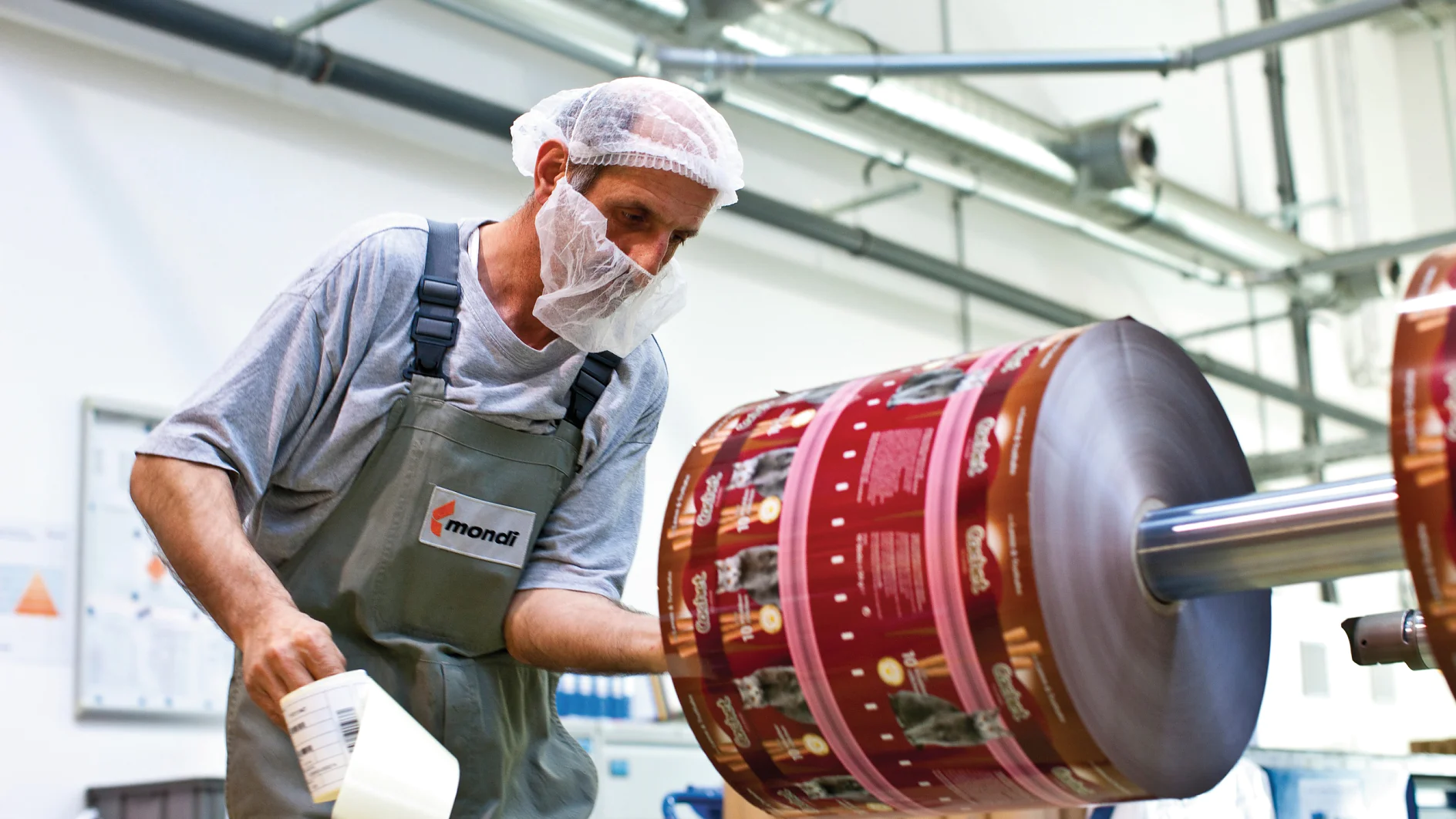 A worker wearing a hairnet and face mask inspects a large roll of printed packaging material in a factory setting. He holds a tool and is dressed in a gray shirt and green overalls. Machinery, tesa tape, and industrial elements are visible in the background. (This text has been generated by AI)