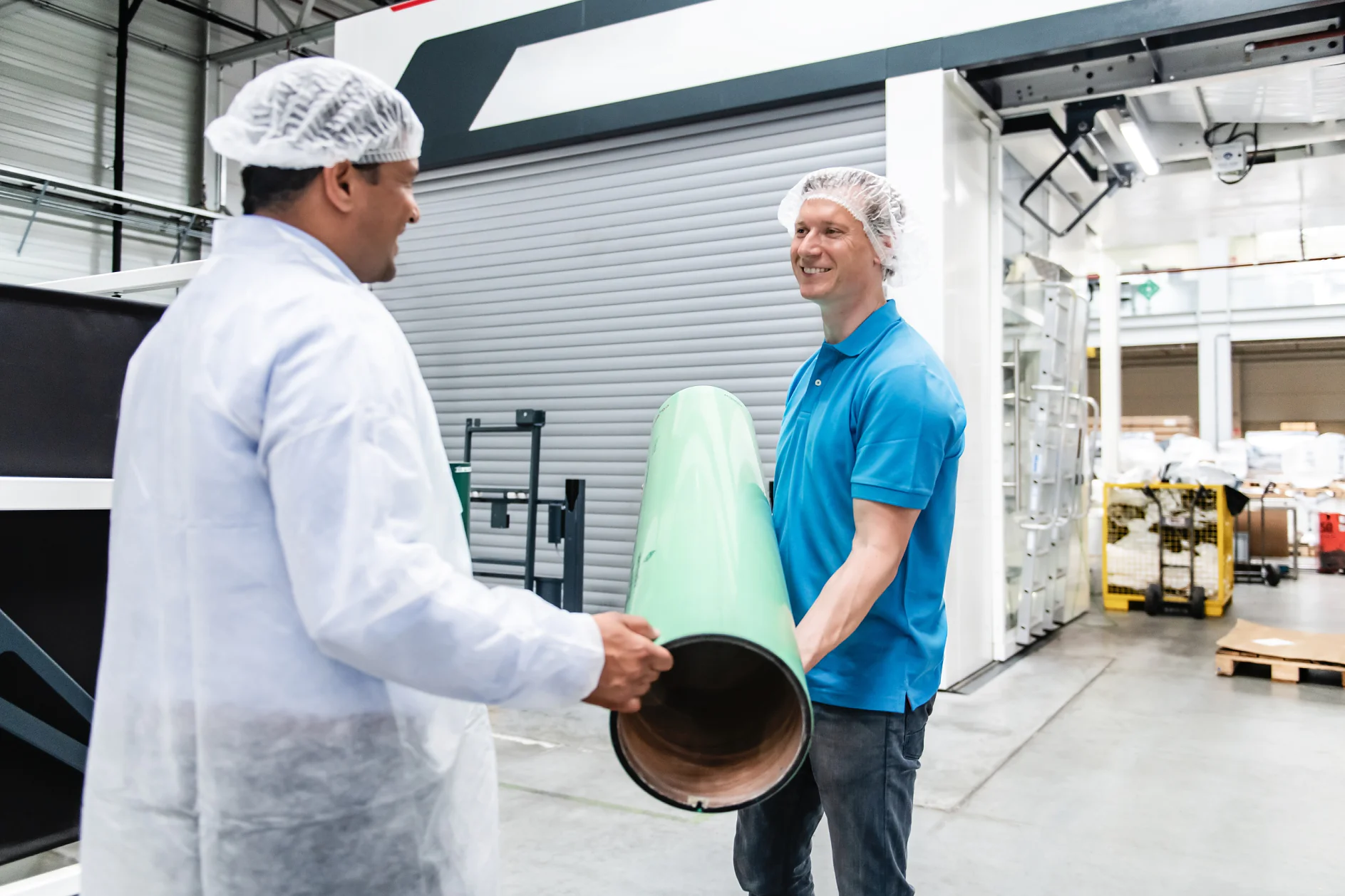 Two men wearing hairnets are in a manufacturing facility. One, dressed in a blue polo shirt, is holding a large green cylindrical object, while the other man, in a white lab coat, observes. Machinery and equipment can be seen in the background. (This text has been generated by AI)