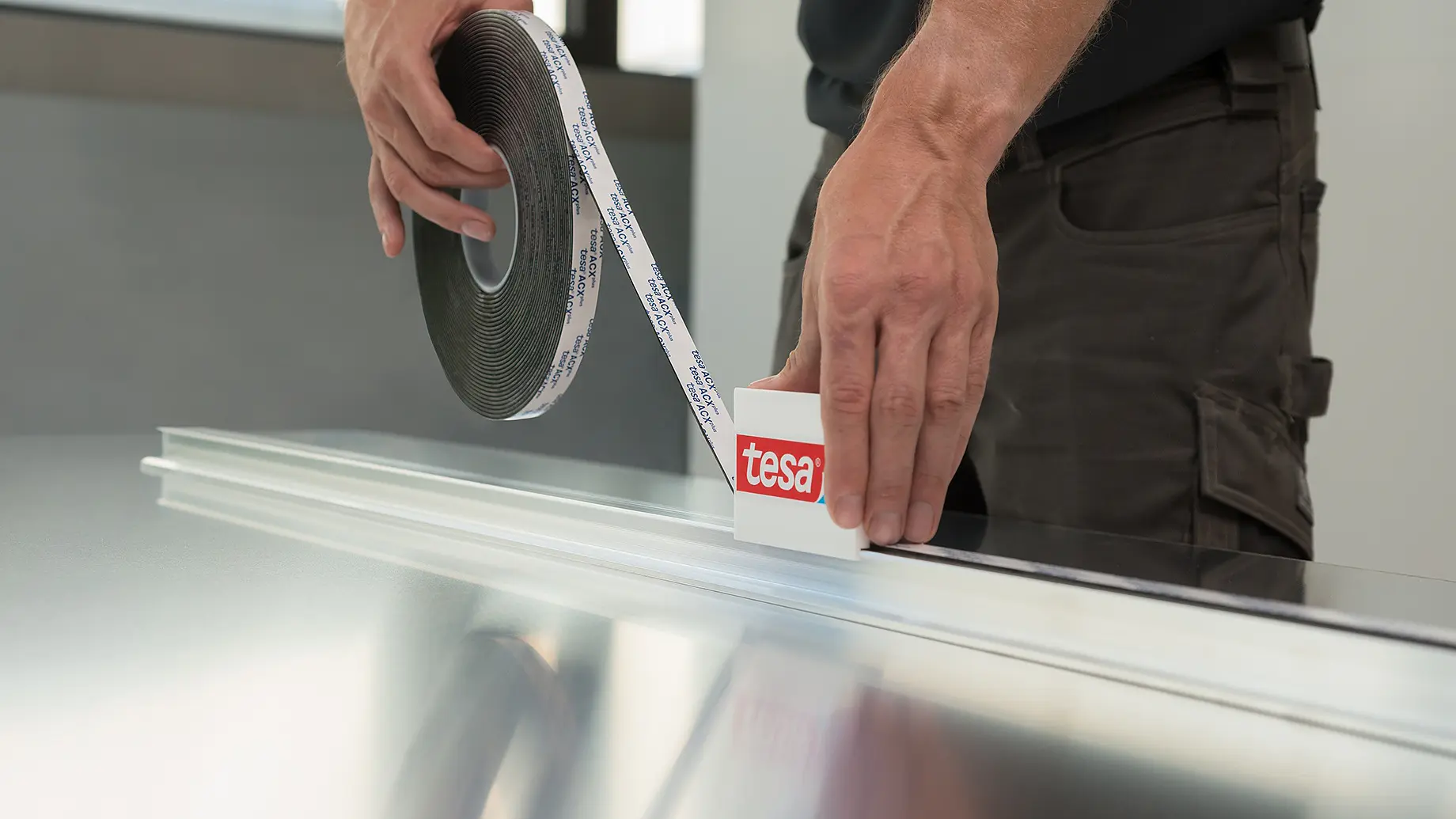 A person applies tesa tape from a roll onto a long, flat surface. The tesa tape roll has a label with the word tesa. The person is wearing a dark shirt and brown pants, and the surface appears to be metallic.