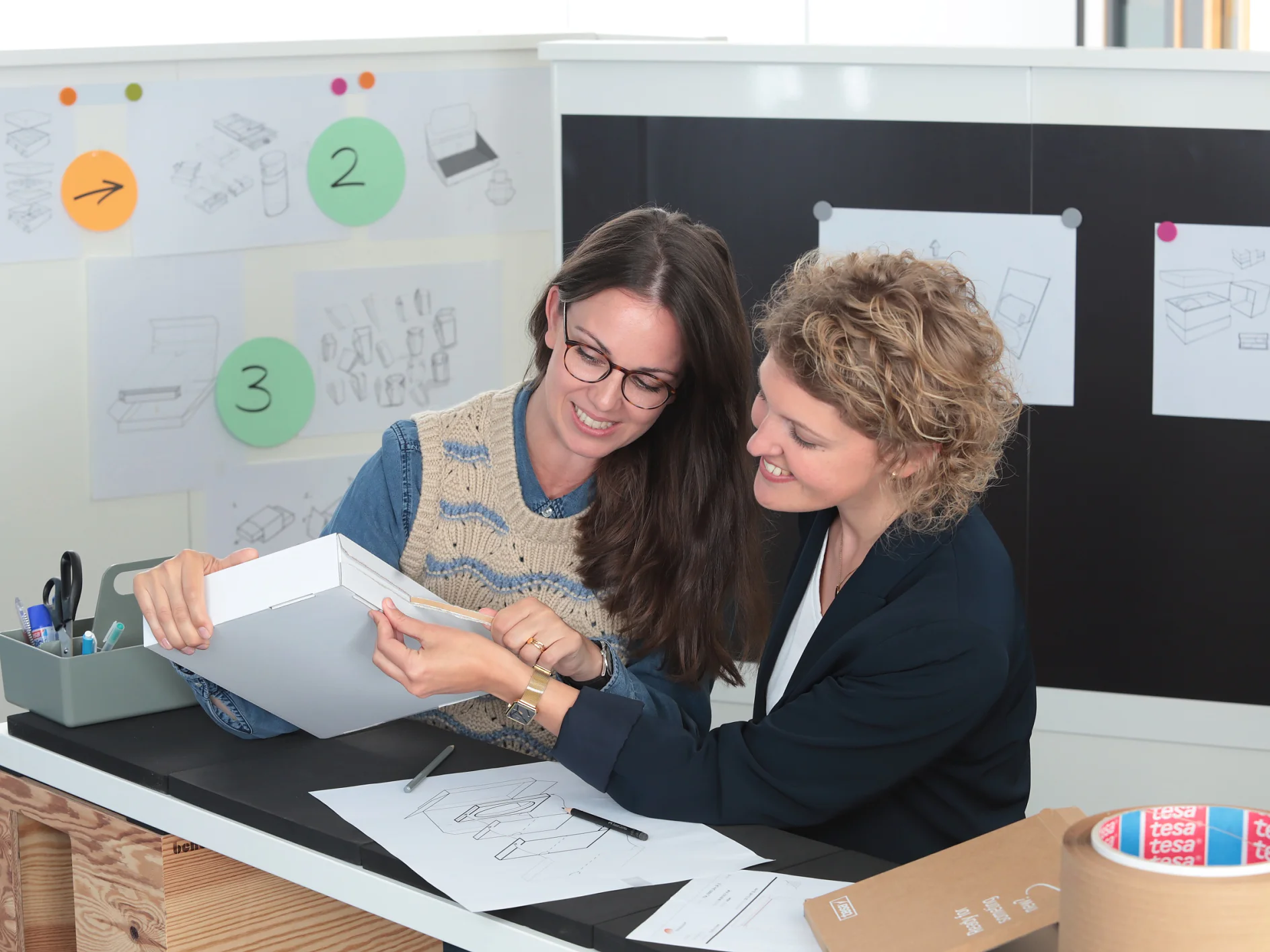 Two people are seated at a table with design sketches, examining a white box. The workspace has sketches and notes on the wall. Various office supplies, including tesa tape and markers, are on the table. Both appear engaged in discussion. (This text has been generated by AI)