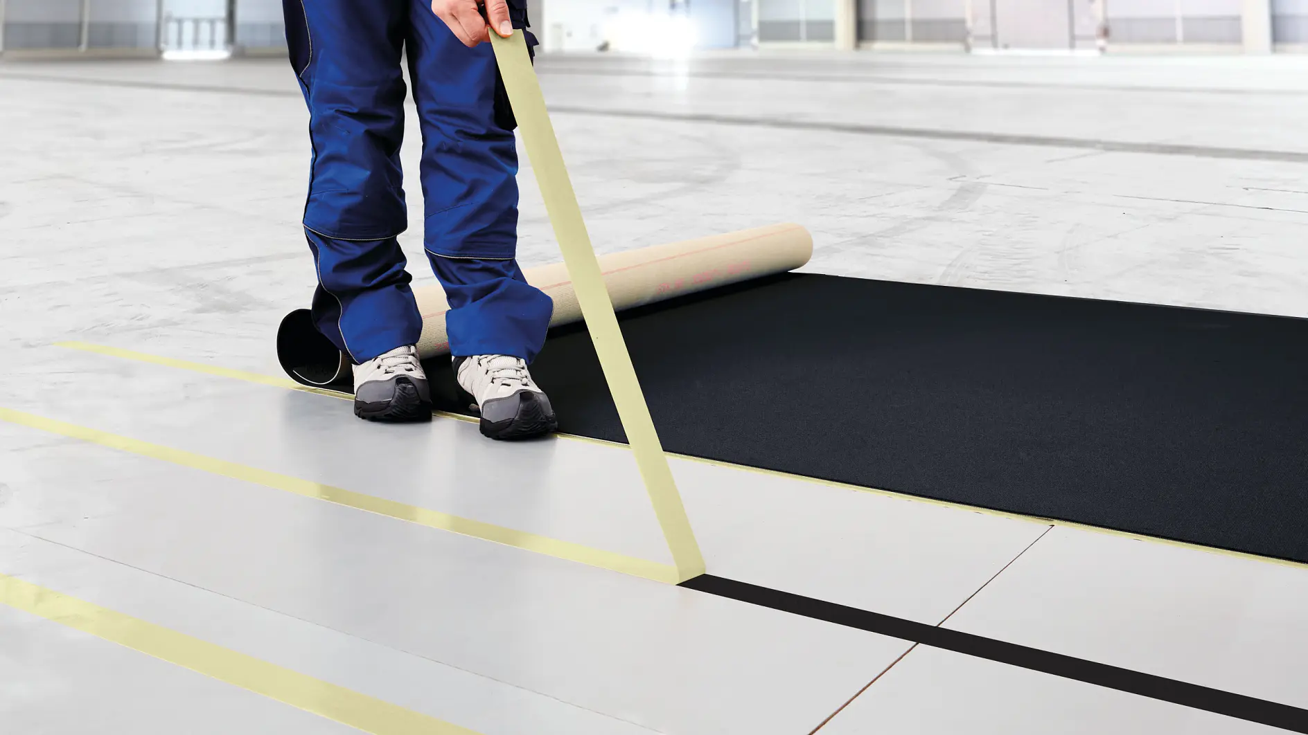 Person unrolling blue paper on a gray floor while applying yellow tape to create parallel lines, illustrating flooring adhesives used for layout marking and precise floor installation preparation.