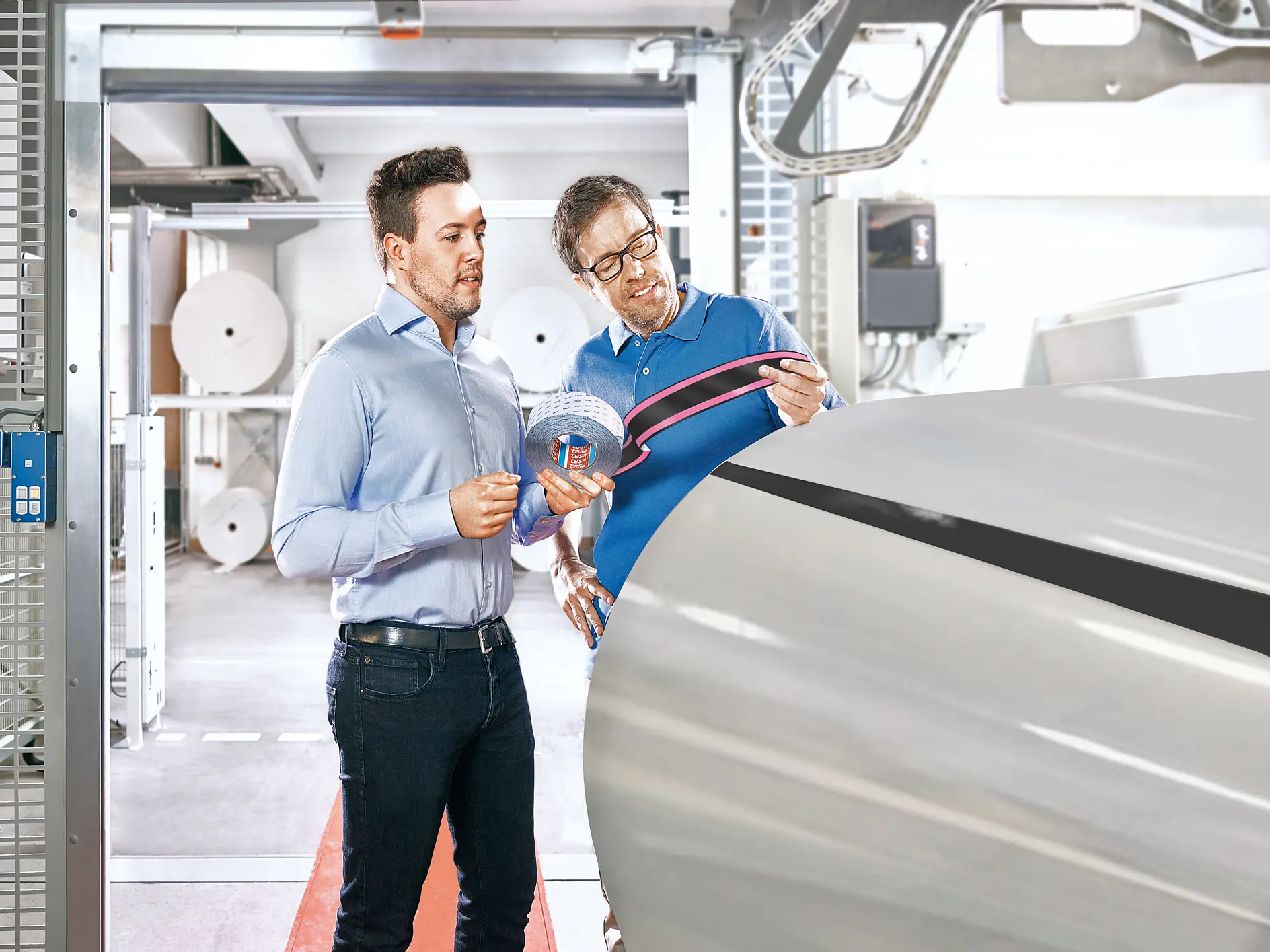 Two men in a manufacturing setting discuss as one holds a roll of printed material and the other points towards a large machine. They appear to be inspecting or analyzing the production process. Rolls of tesa tape can be seen in the background.