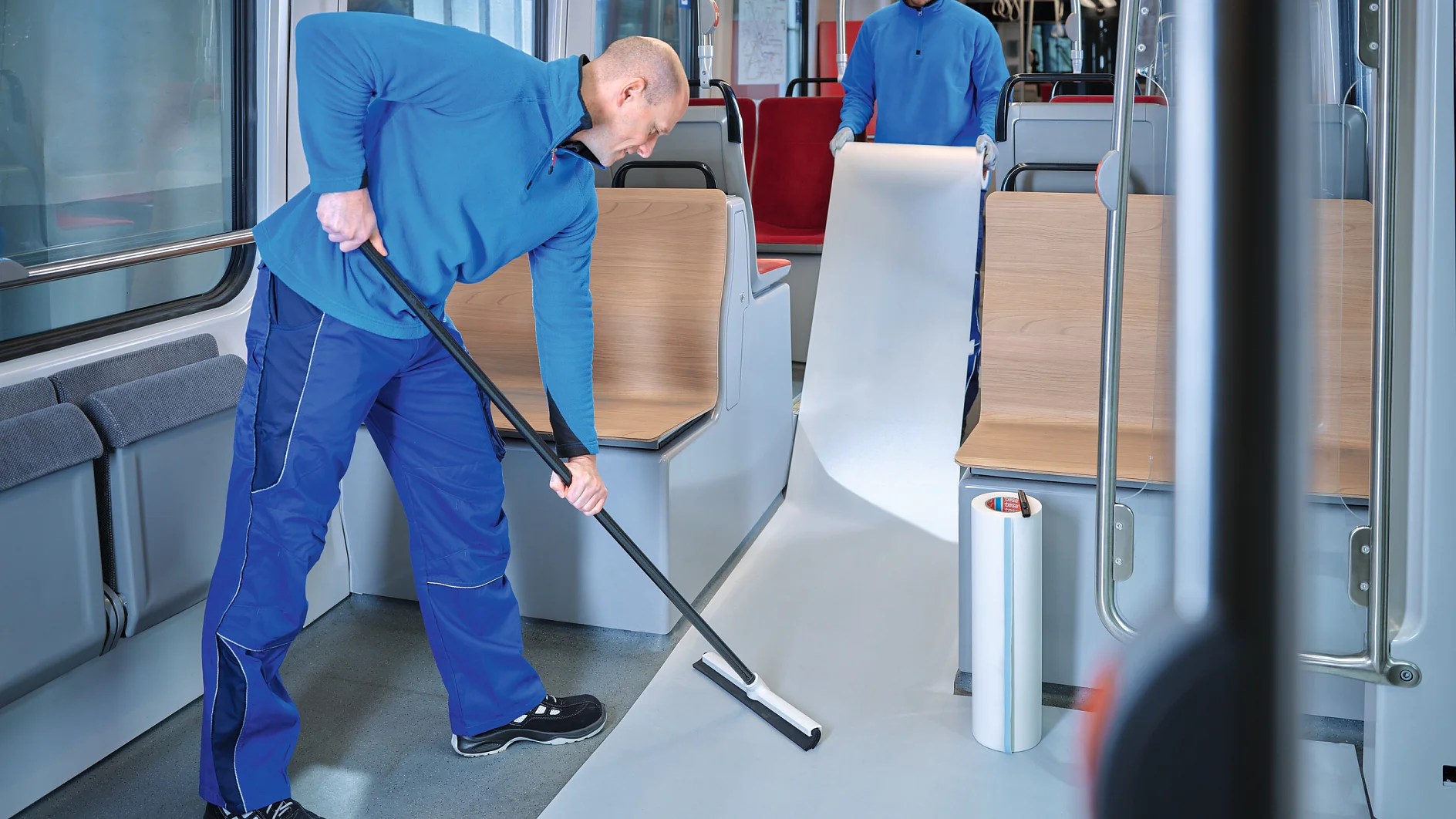 two workers in blue uniforms installing white flooring inside a modern public transport vehicle, one rolling out material while the other smooths it