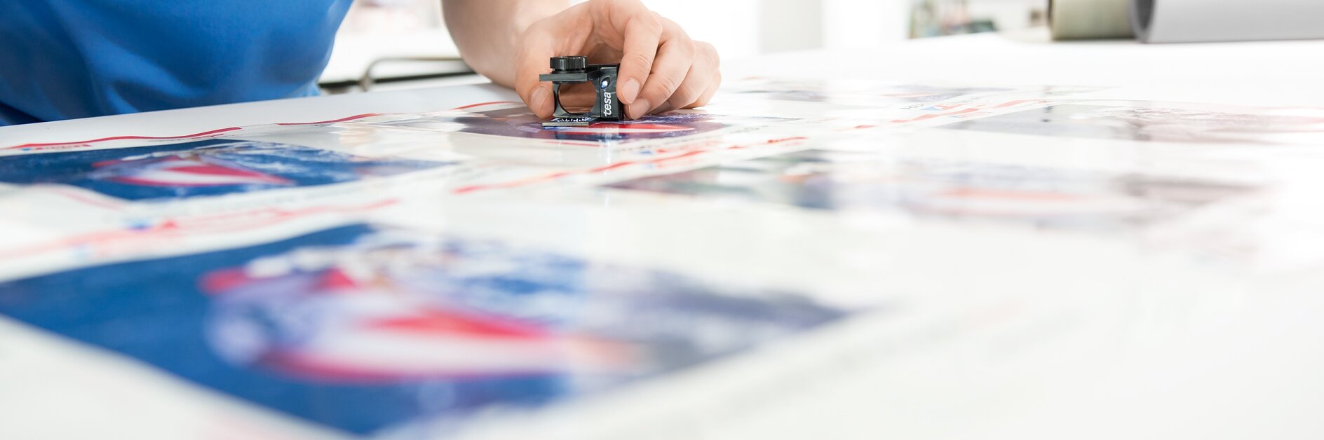 A person in a blue polo shirt is inspecting printed materials on a table using a magnifying tool. The shirt has a red logo. The workspace has bright lighting, and there are blurred images in the background. tesa tape is visible on the edge of the table, ready for use. (This text has been generated by AI)