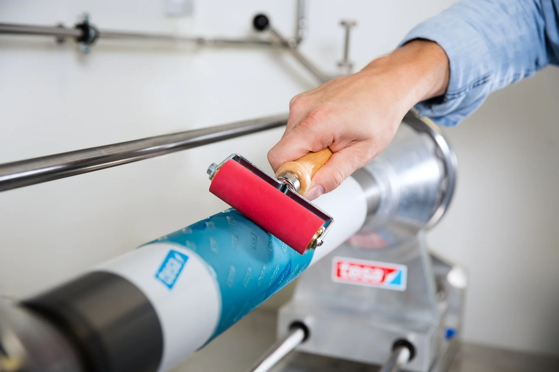 FP&P_Flexographic-Printing_Plate-Mounting-072-Screen A person uses a red roller on blue tesa tape wrapped around a cylindrical machine part. The setup includes stainless steel components and visible labels. The person is wearing a light blue sleeve. (This text has been generated by AI)