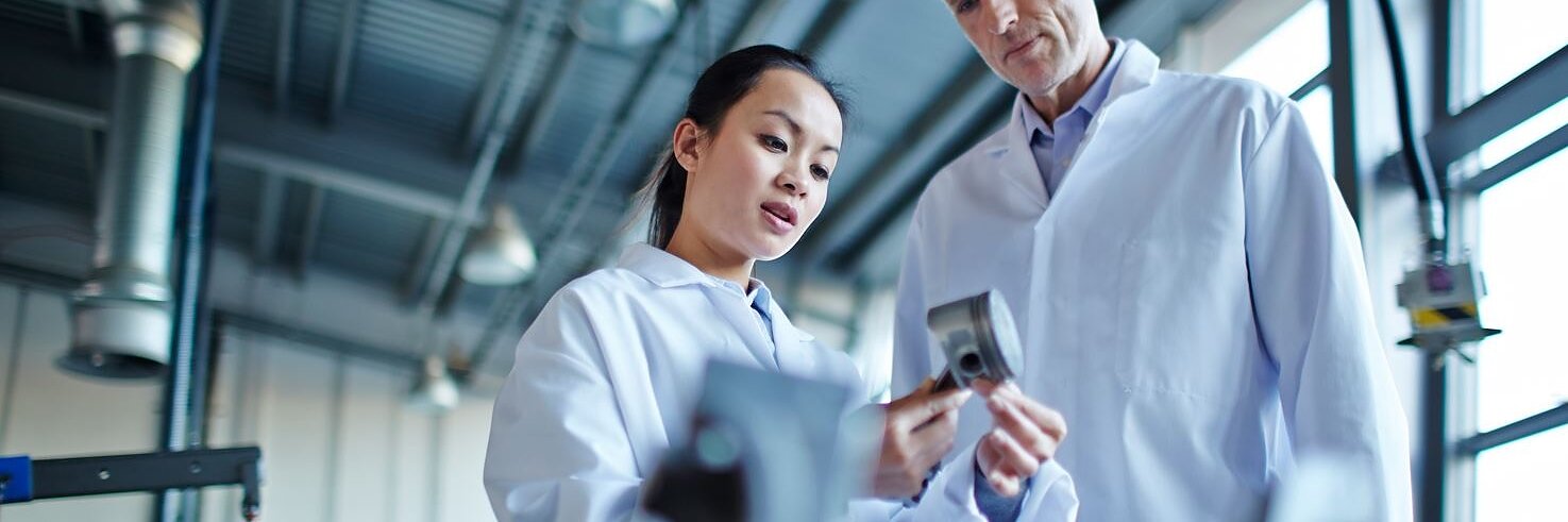 Two people in lab coats examine a metallic object in a bright industrial setting. The person on the left is pointing at the object, while the person on the right observes. Large windows and machinery are visible in the background, with tesa tape used prominently within the space for various applications. (This text has been generated by AI)