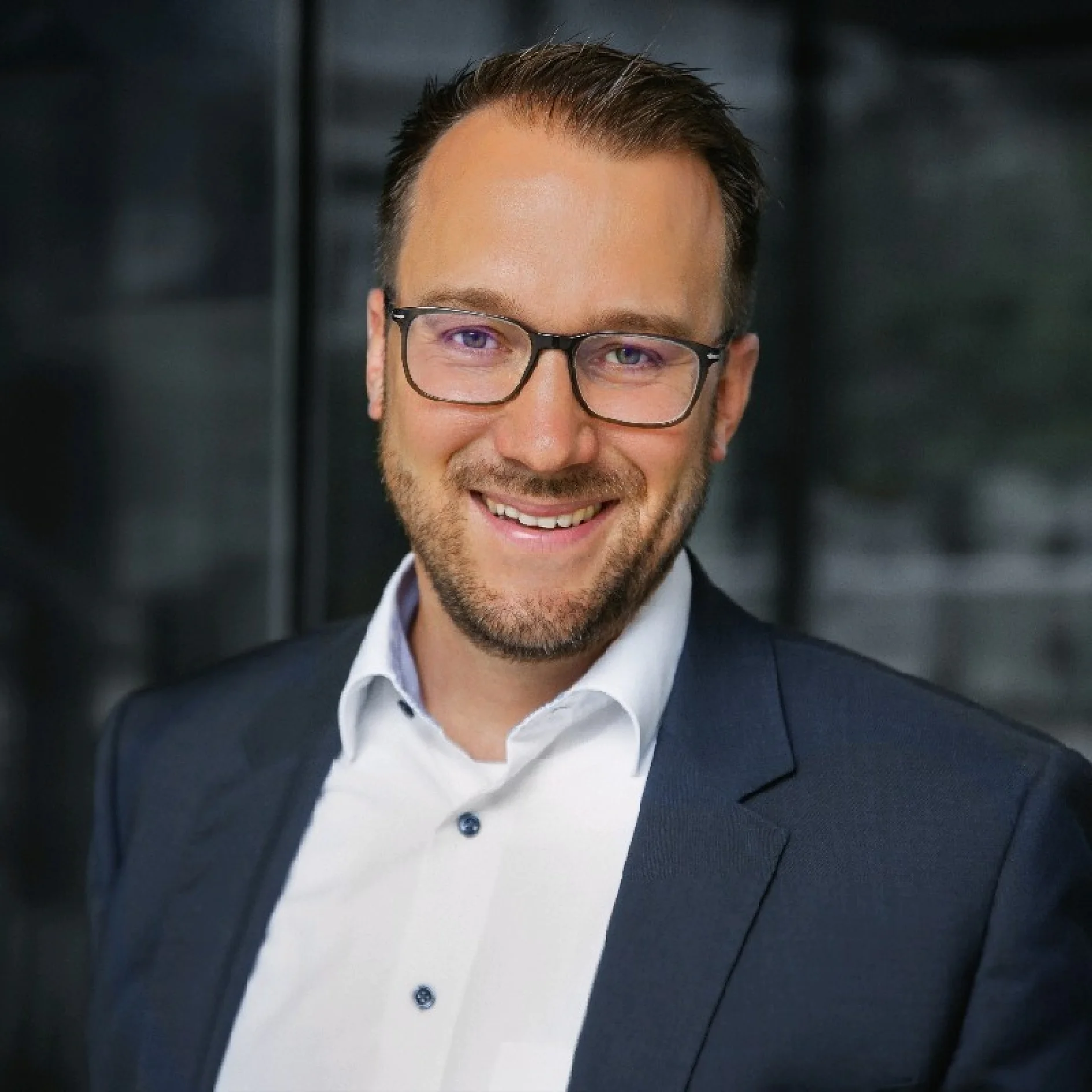 A man with short hair and glasses is smiling at the camera. He is wearing a dark suit jacket over a white shirt in an indoor setting with a dark, blurred background. (This text has been generated by AI)