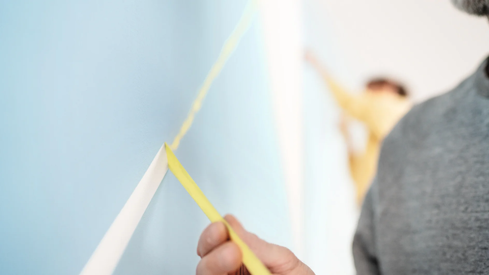 A close-up of two people applying yellow tesa tape in a diagonal line on a light blue wall. The person in the foreground is in focus, wearing a gray sweater, while the other person in the background is blurred. (This text has been generated by AI)