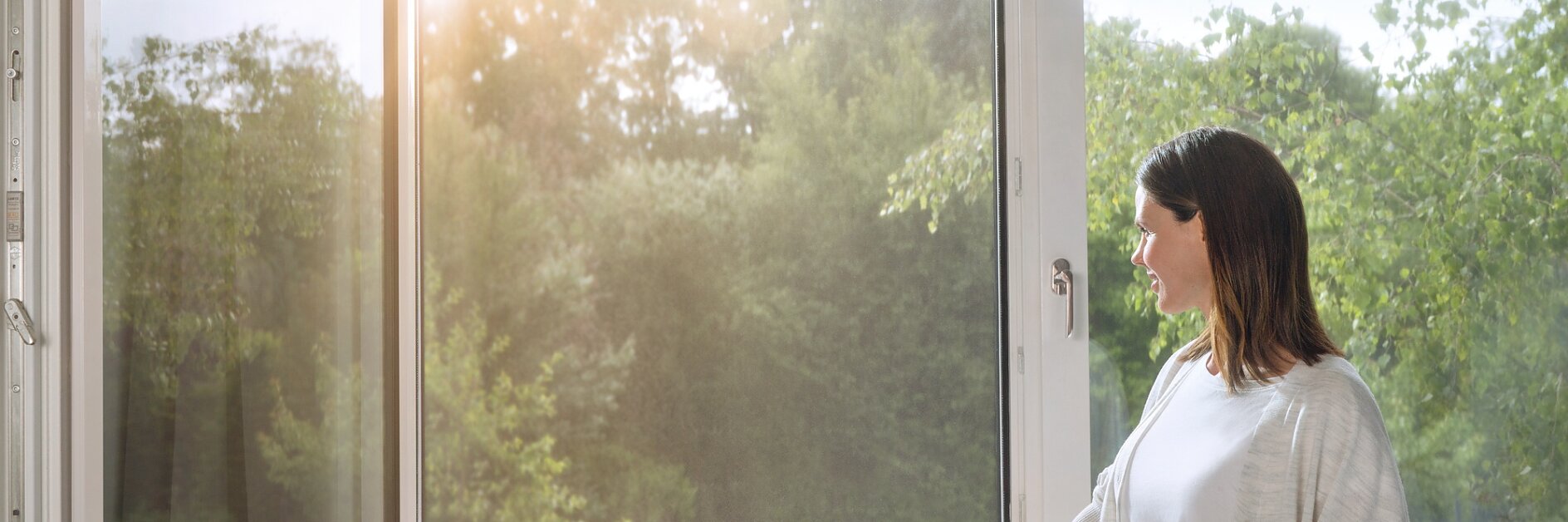 A woman stands by an open window, looking outside. Sunlight filters through, illuminating the greenery of trees in the background. She is wearing a light cardigan and jeans, with one hand resting on the windowsill secured with tesa tape. (This text has been generated by AI)