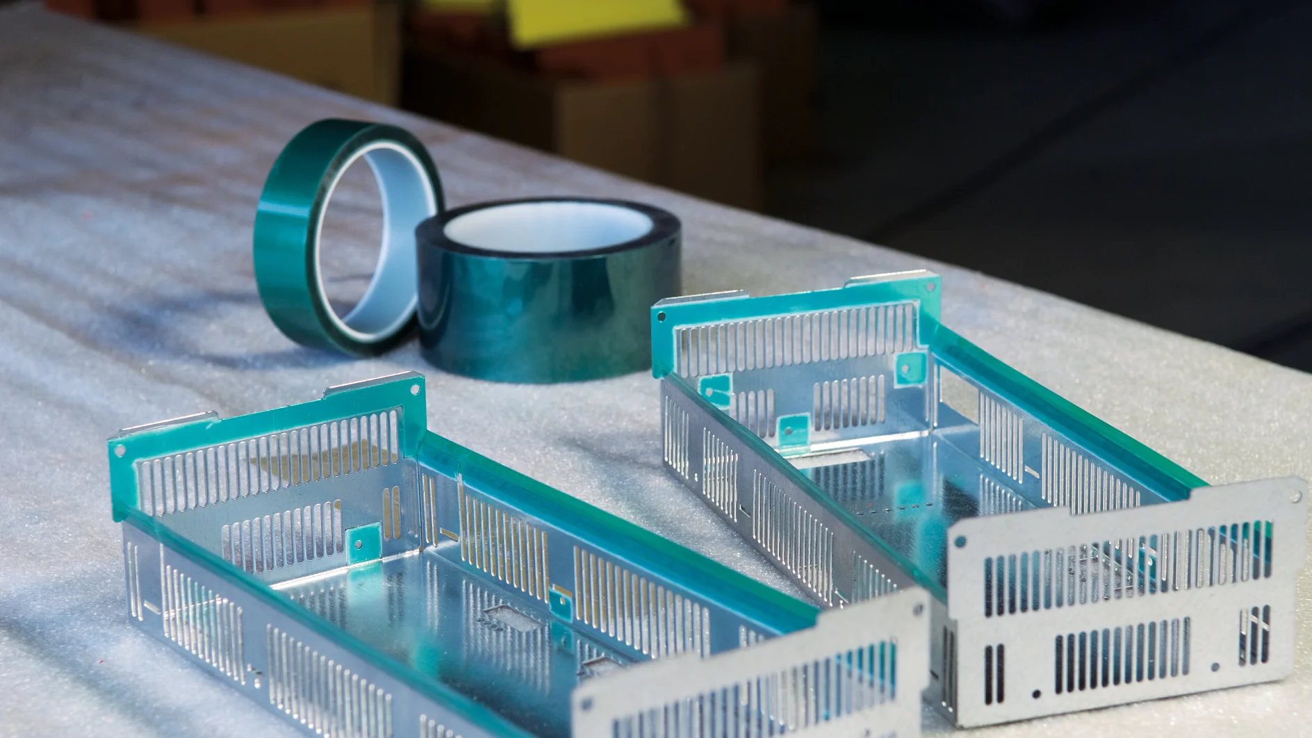 tesa-50600-polyester-silicone-masking-tape-green-translucent-ap-001.tif-cms Two rectangular metal frames with slots are on a textured surface. Behind them are two rolls of green tesa tape. The background is out of focus, showing stacks of paper or cardboard. (This text has been generated by AI)