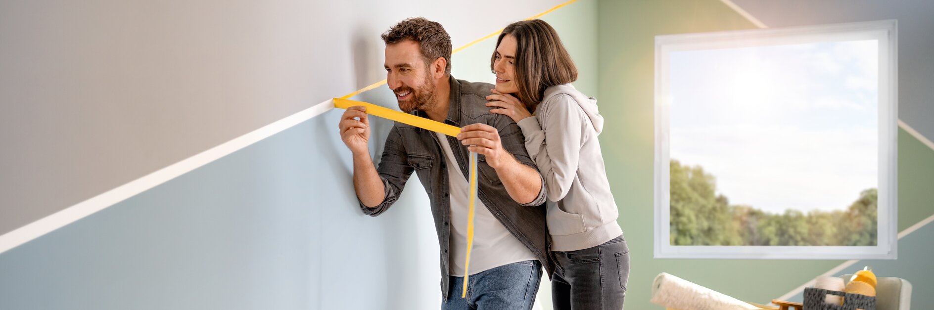 A man is holding a measuring tesa tape against a wall with geometric patterns. A woman stands close behind him, assisting with the tesa tape. The room contains a chair and a basket near a large window, showcasing greenery outside. They seem to be decorating the space. (This text has been generated by AI)