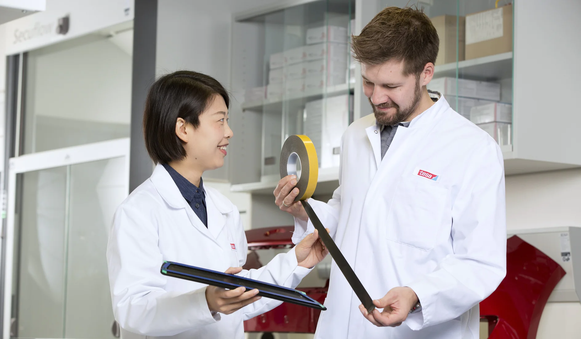 Two people in lab coats are standing in a laboratory. One person holds a tablet and smiles, while the other is unwinding a roll of tesa tape. Shelves with boxes and equipment are visible in the background. (This text has been generated by AI)