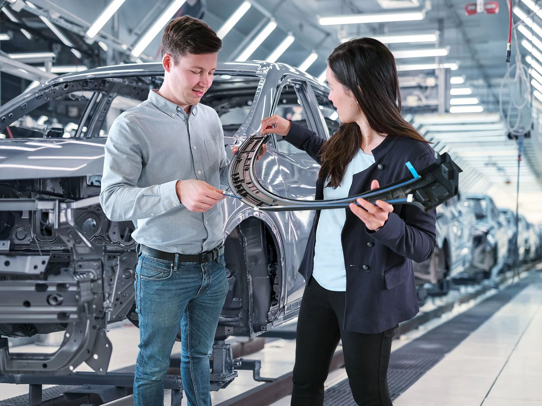 Two people are examining a car part inside an automotive factory. The man holds a segment of the part, while the woman holds the other part, and they converse. Car skeletons are visible in the background under bright lighting, with rolls of tesa tape nearby on workbenches for assembly use. (This text has been generated by AI)