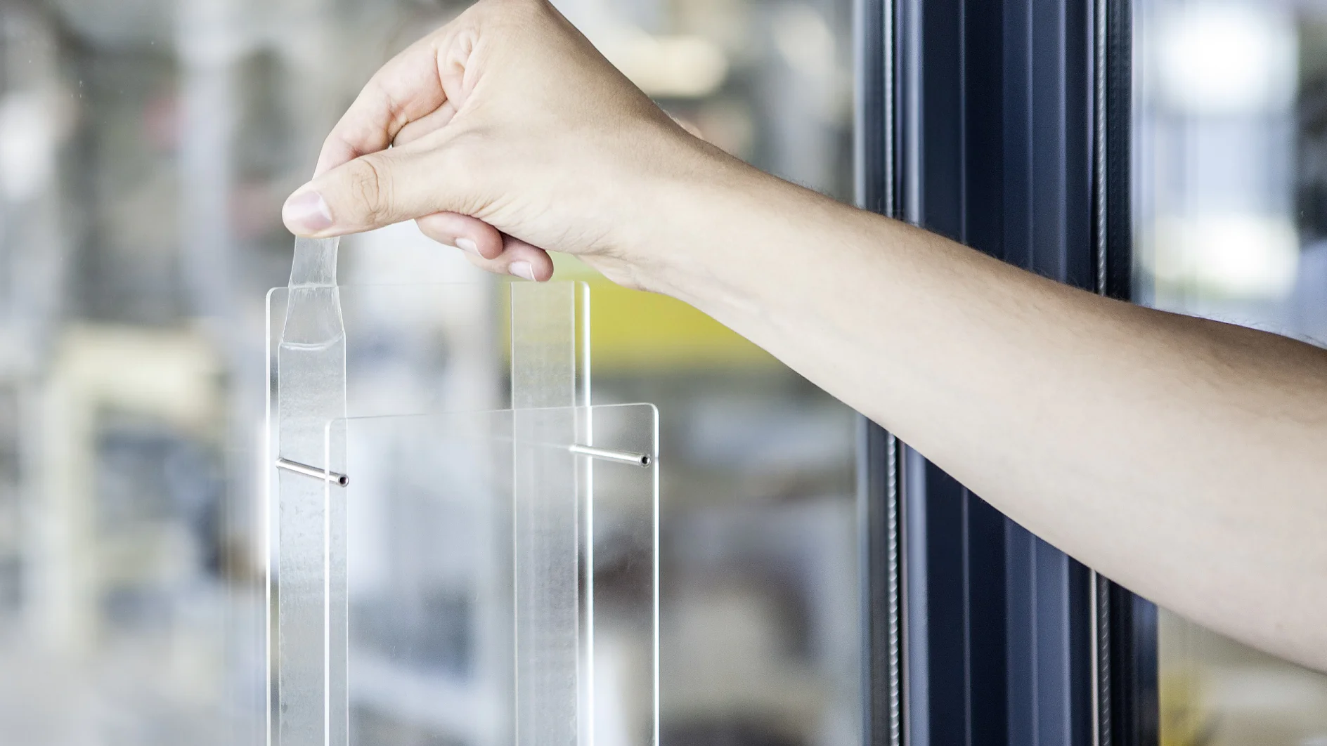 bild-entfernbare-klebebaender-2-300dpi A persons hand is seen peeling off a protective film from a transparent glass or acrylic surface, using tesa tape. The background is out of focus with hints of an indoor setting. (This text has been generated by AI)