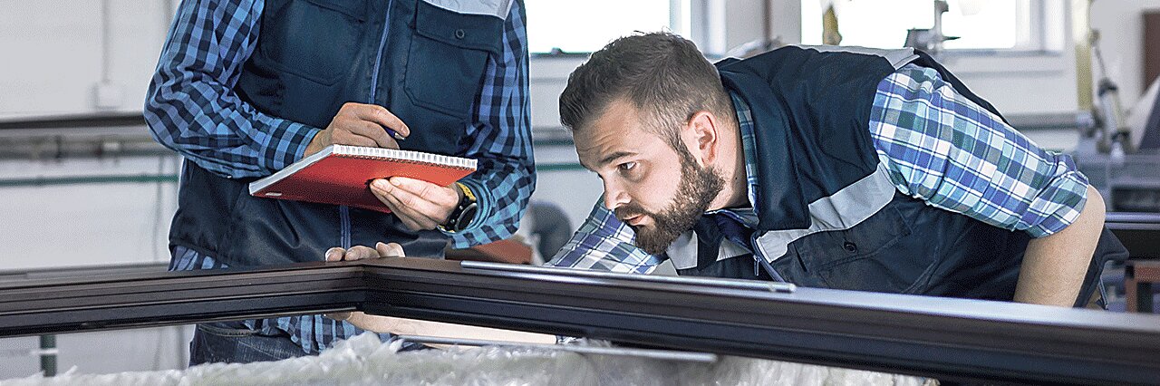 Two men wearing checkered shirts and vests are in an industrial setting. One is inspecting a metal frame while the other takes notes in a red notebook. The background shows various equipment and work materials, including rolls of tesa tape. (This text has been generated by AI)