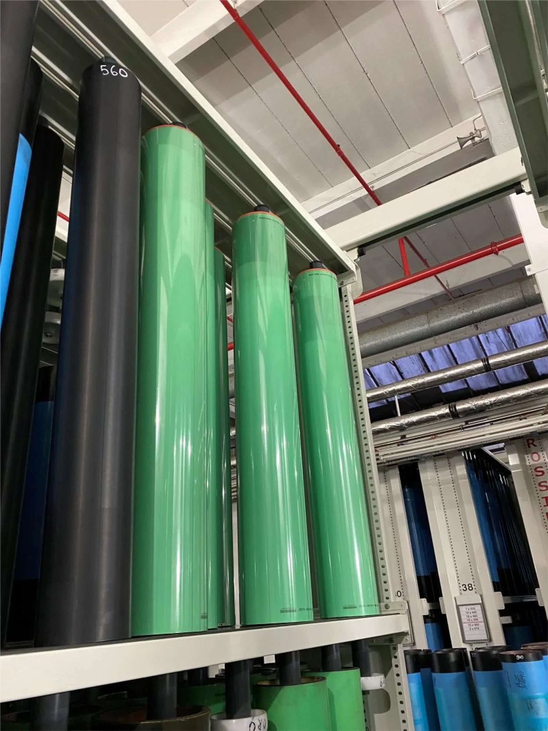 Several large green and black cylindrical rolls of tesa tape are stored upright on metal shelves in an industrial setting. Red pipes and a metal ceiling structure are visible above the shelves. (This text has been generated by AI)