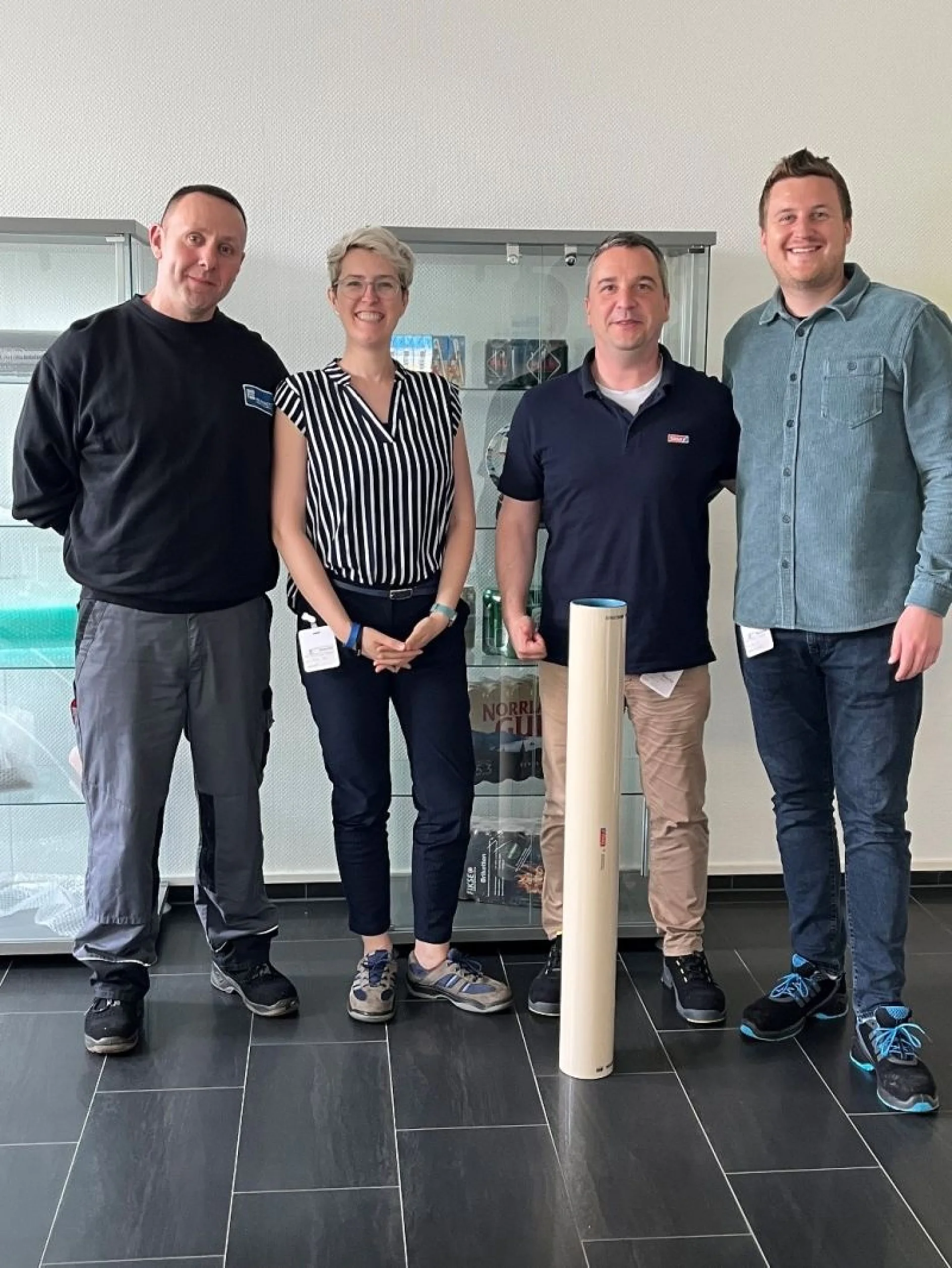 Four people stand indoors on a dark tile floor. Three men and one woman pose with a roll of tesa tape on the floor. A display cabinet is visible in the background. They are casually dressed and smiling. (This text has been generated by AI)