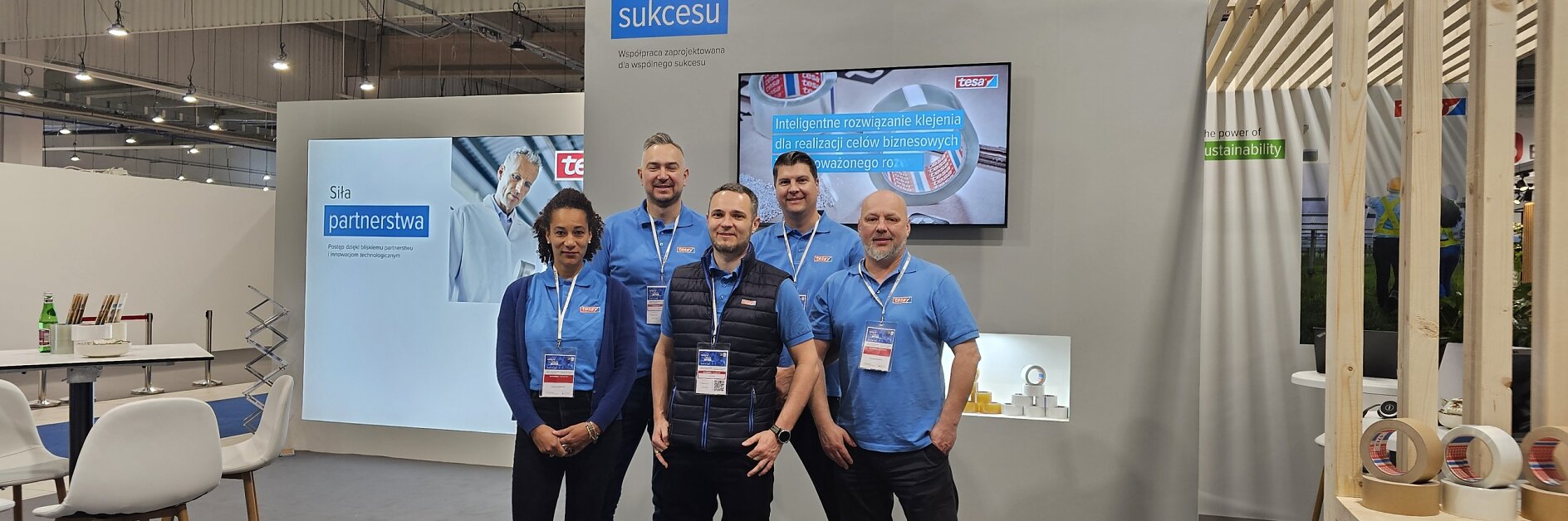 Five people stand in front of a booth at an exhibition hall. They are wearing casual blue shirts with event badges. The booth displays the tesa tape logo and promotional materials. A table with chairs is visible on the left. (This text has been generated by AI)