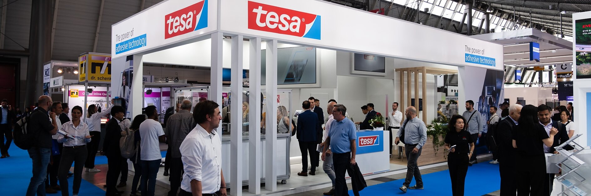 People are gathered around a trade show booth with a prominent tesa logo. The booth has blue carpeting and white panels. Attendees are walking, browsing displays, and engaging in conversations. A large overhead sign reads The future of converting & tesa automation. (This text has been generated by AI)