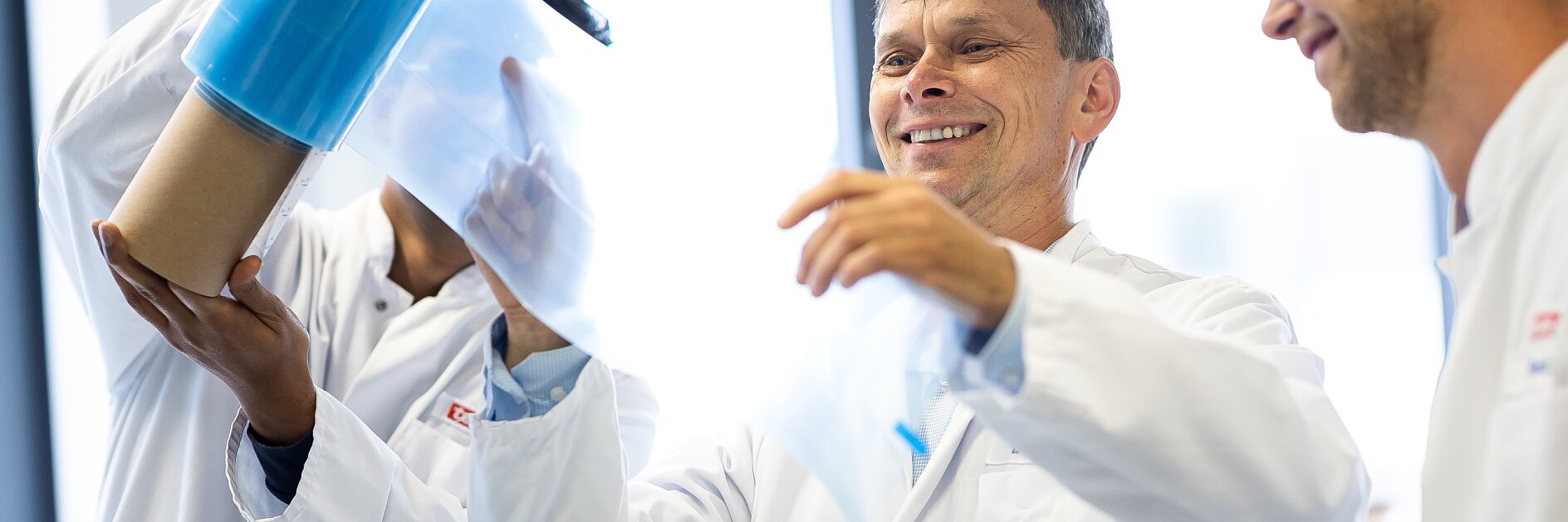 Three people in lab coats examine a roll of transparent blue tesa tape in a bright laboratory setting. One person holds the tesa tape upright, while another pulls a section away. They appear focused and engaged.