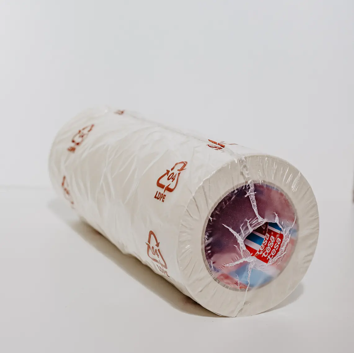 A roll of white plastic wrap is standing upright against a plain white background. The packaging is partially transparent, revealing the inner cardboard tube and multicolored label. Red recycling symbols and text can be seen on the plastic.