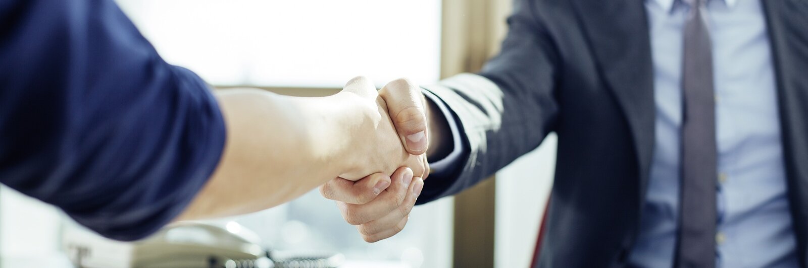 Two people are shaking hands across a desk in a brightly lit office. One person is wearing a suit, while the other has a casual shirt. A tablet and some papers with graphs are on the desk, along with some tesa tape. (This text has been generated by AI)