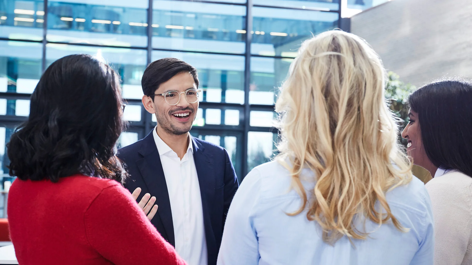 tesa_HR_employees_05_72dpi-cms A man in a suit and glasses is smiling while talking to three women, one in a red sweater and two with light-colored tops. They are standing in a modern office with large windows in the background, with rolls of tesa tape placed neatly on the desk nearby. (This text has been generated by AI)
