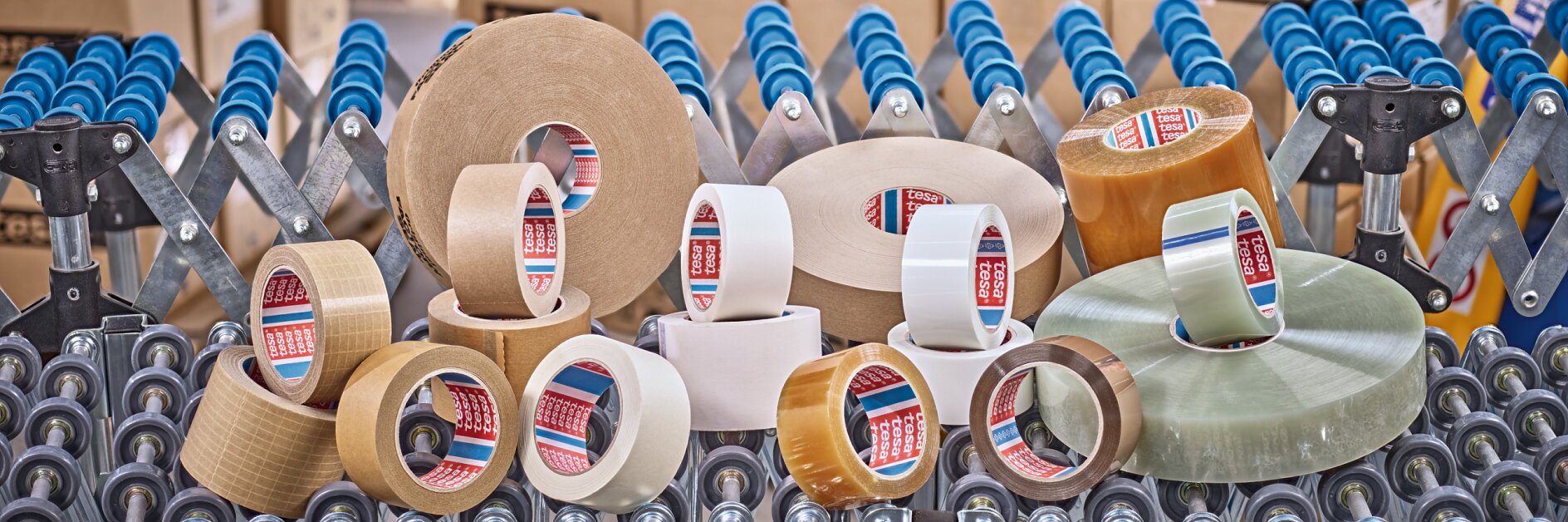 Various rolls of adhesive packaging tesa tape are displayed on a roller conveyor. The tesa tapes vary in color, including clear, brown, and beige. In the background, there are stacks of cardboard boxes and blue roller tracks. (This text has been generated by AI)
