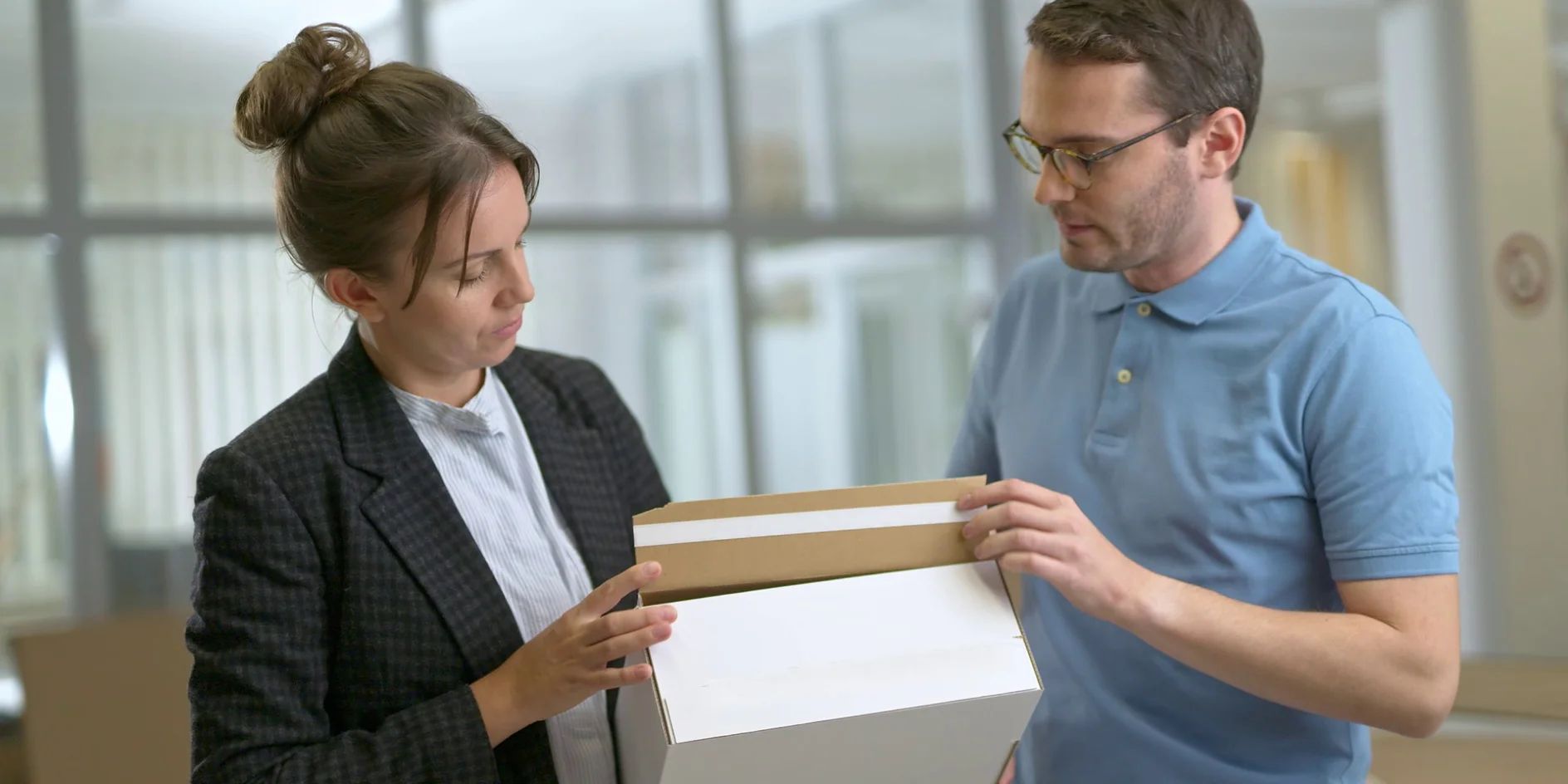 A man in a blue polo shirt and glasses is holding a white cardboard box secured with tesa tape, while a woman in a checkered blazer is looking at it. They are standing in an office setting with glass partitions in the background. (This text has been generated by AI)