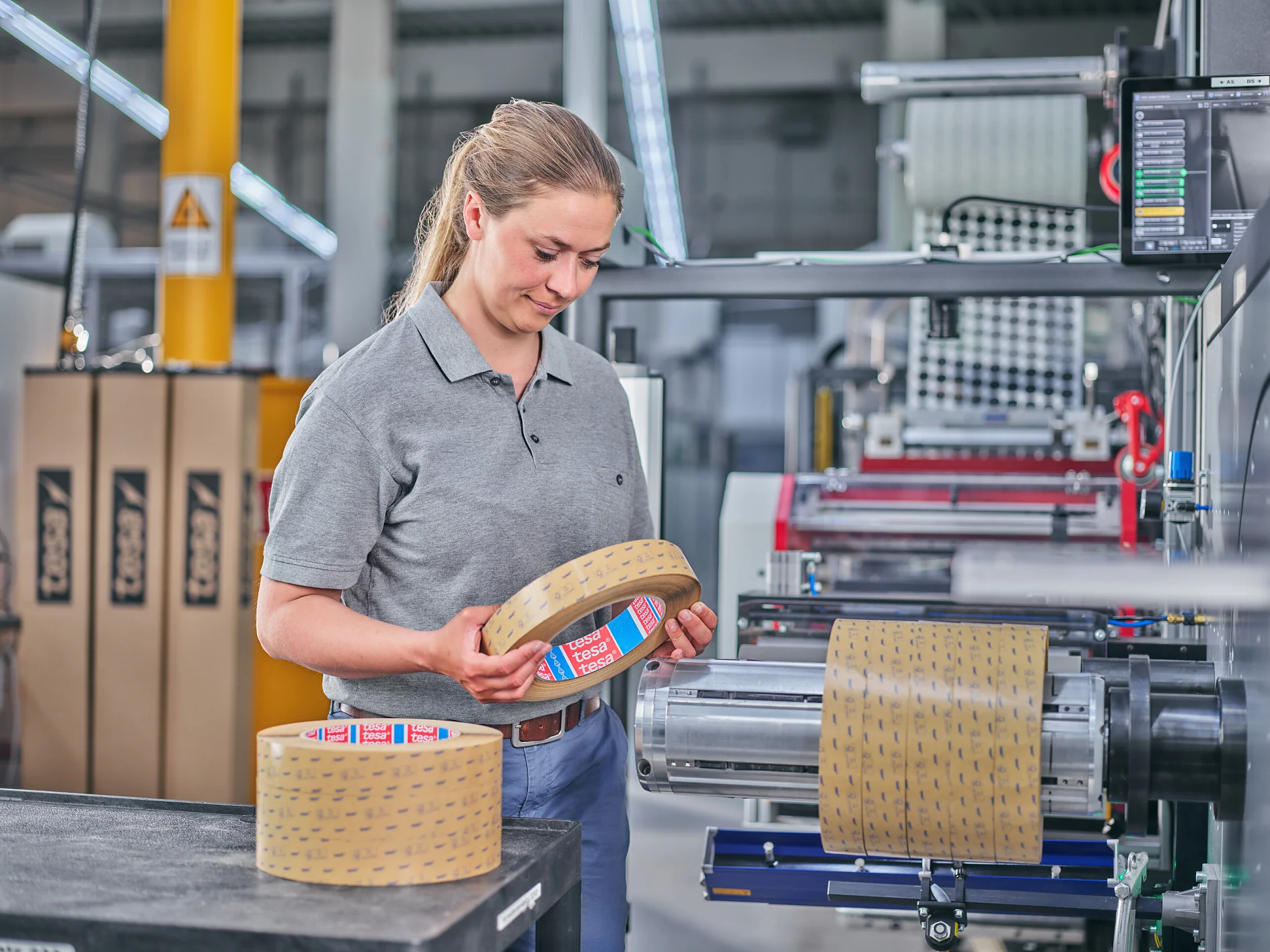 worker in gray tesa shirt handling adhesive tape rolls in an industrial packaging facility with machinery and stacked boxes nearby