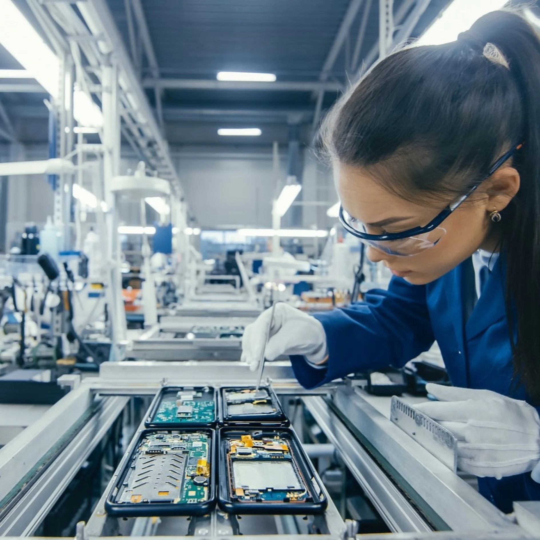 A woman in a blue lab coat and safety glasses examines disassembled smartphone components on a conveyor belt in a factory setting. She holds a tool and wears gloves, indicating a focus on detail and safety in the work environment. All the components are secured with tesa tape for precision assembly. (This text has been generated by AI)