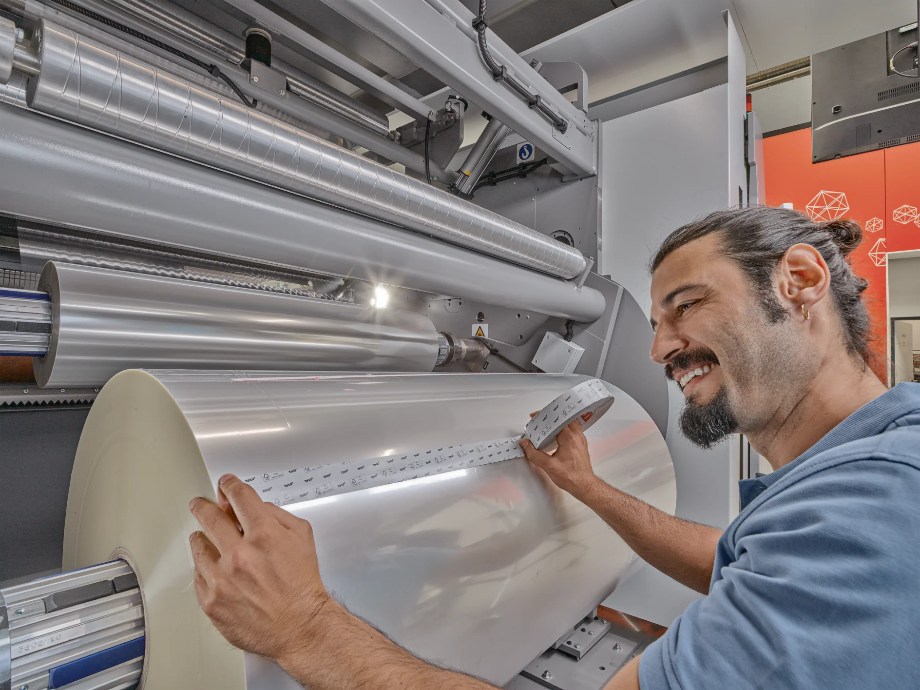 A man with a beard and a ponytail operates a machine, handling a large roll of reflective material, possibly for printing. He is pressing a small strip of tesa tape inside the machine, which is surrounded by metallic rollers. (This text has been generated by AI)