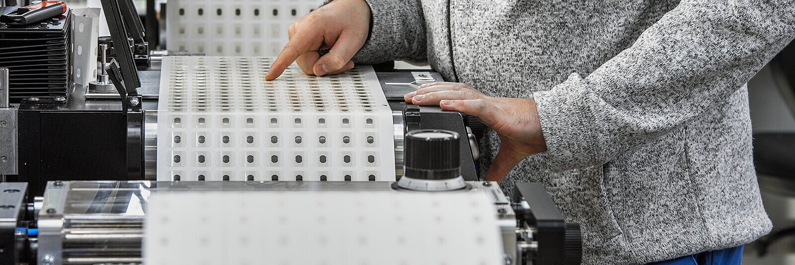 A person operates machinery producing tesa tape rolls. The individual is wearing a gray sweater with a logo and is adjusting the settings using a control knob. The tesa tape on the machine has a white background with a pattern of small circles. (This text has been generated by AI)