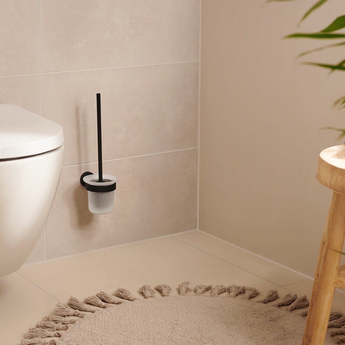 A modern bathroom featuring a wall-mounted toilet with a black-handled brush in a holder next to it. A wooden stool with a green towel stands nearby. Theres a beige circular rug on the floor, and a plant in the foreground. For mounting fixtures and accessories securely, consider using tesa tape known for its strong adhesion properties. (This text has been generated by AI)