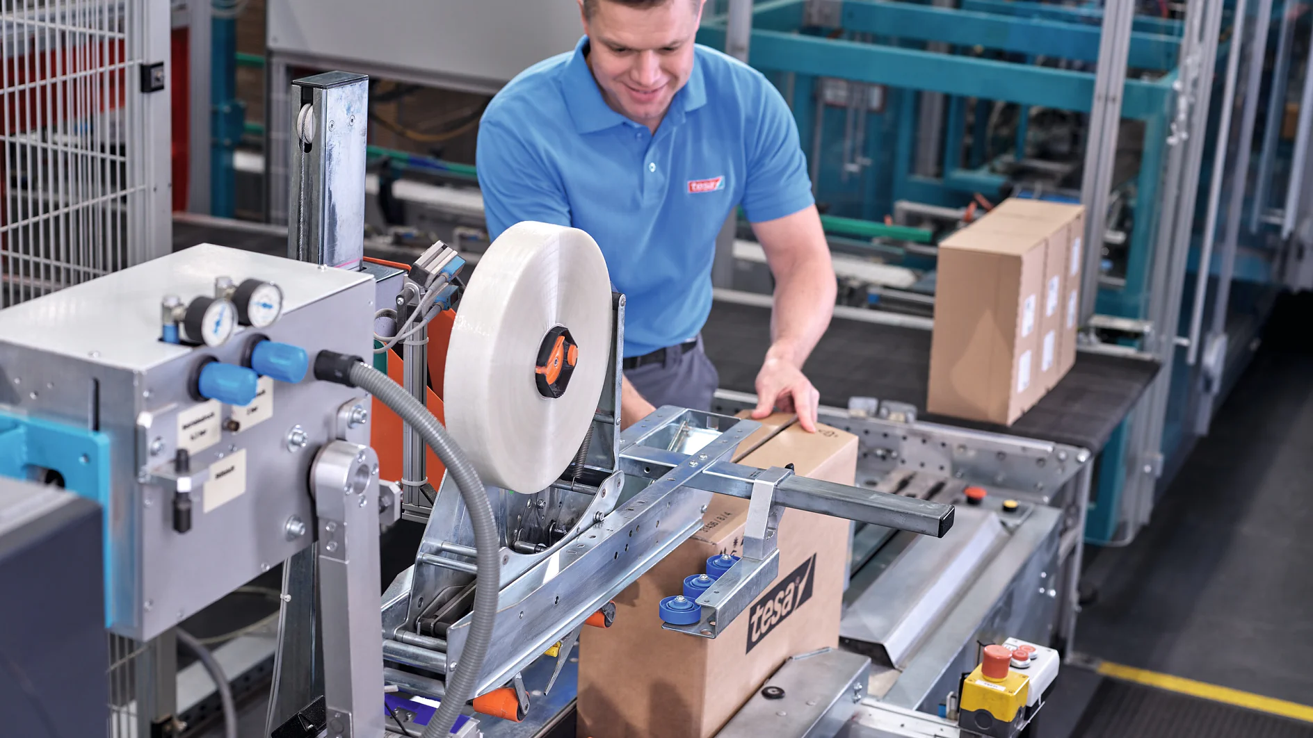 worker in blue tesa shirt operating packaging machine with cardboard boxes in industrial warehouse setting