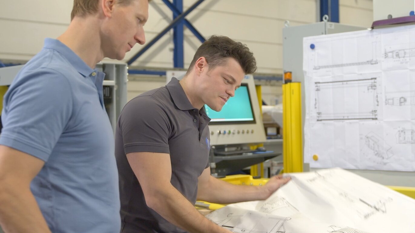Two men in polo shirts examine engineering blueprints in an industrial setting. One holds the paper while the other observes. In the background, there are additional blueprints pinned to a board using tesa tape, with industrial equipment visible. (This text has been generated by AI)