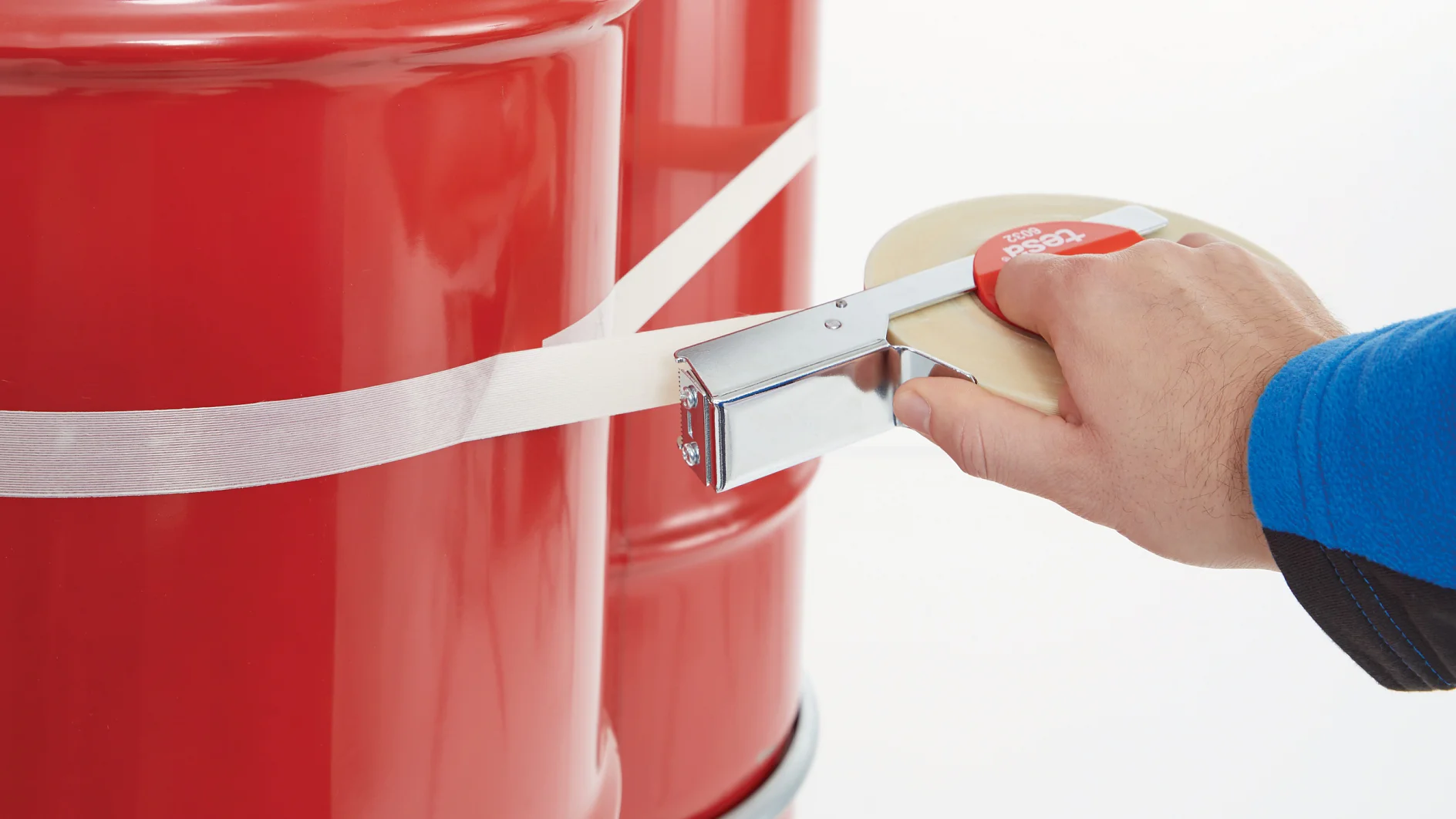 hand using a tesa tape dispenser to apply clear tape around a red metal drum in a bright indoor setting (This text has been generated by AI)