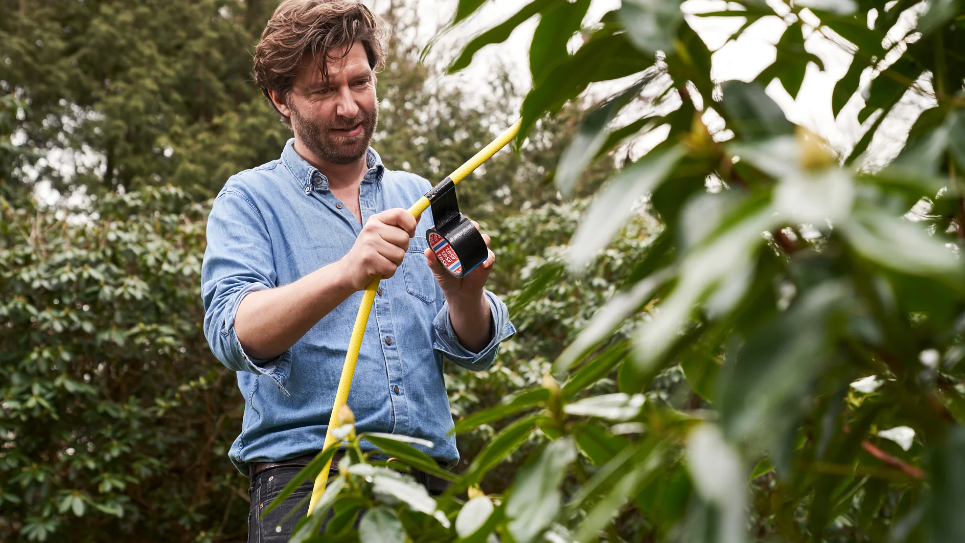 A man wearing a denim shirt is wrapping black tesa tape around a yellow garden hose in a leafy outdoor setting. (This text has been generated by AI)