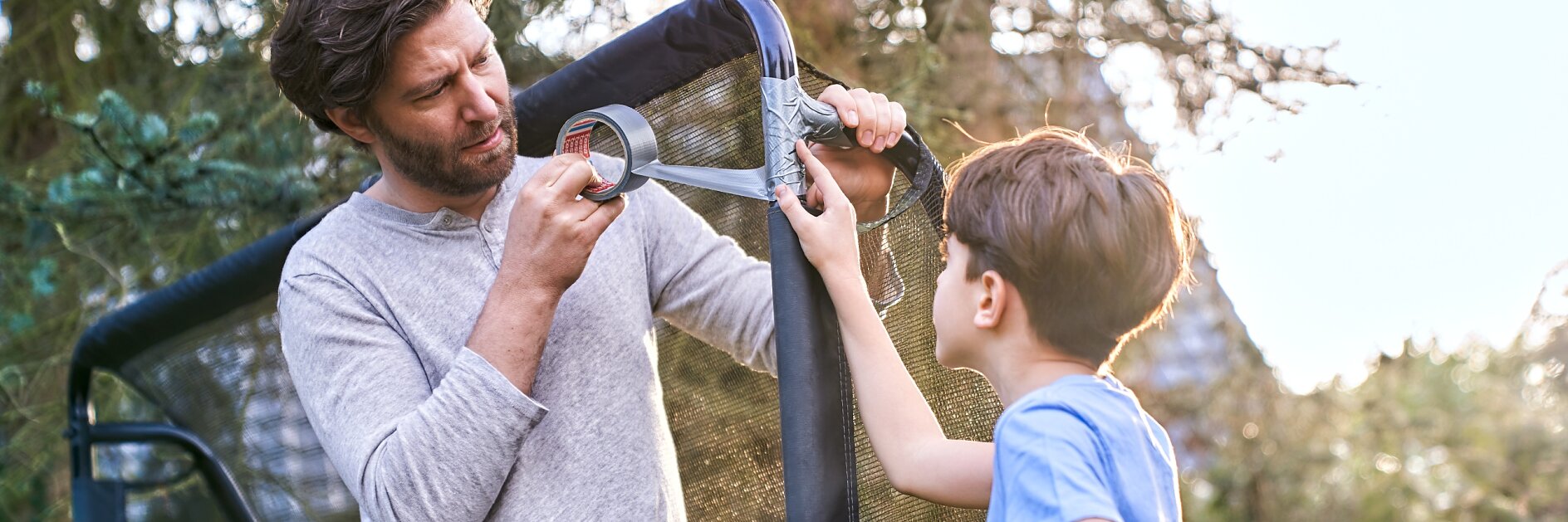 A man is using tesa tape to repair a net structure outdoors while a boy holding a football watches. They are standing near trees, with the sun shining in the background. The scene suggests a collaborative effort. (This text has been generated by AI)